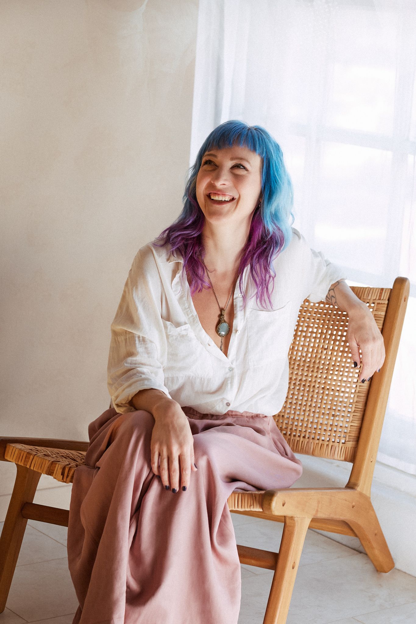 A person with vibrant blue and purple hair laughs while sitting in a wooden cane chair wearing a white shirt and pink pants.