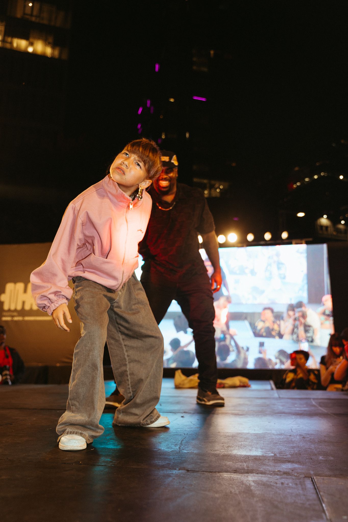Figure in a gray outfit performs a dynamic breakdance move on a dark stage with bright lights in the background.