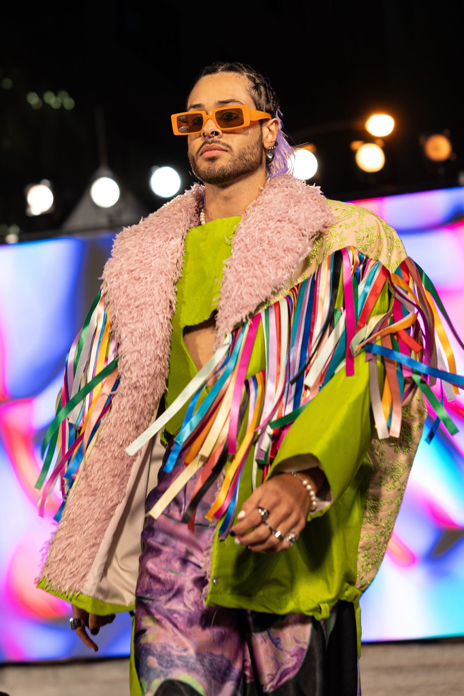 Performer wearing colorful striped jacket, pink fur collar, lime green top and orange sunglasses on stage with blurred lights.