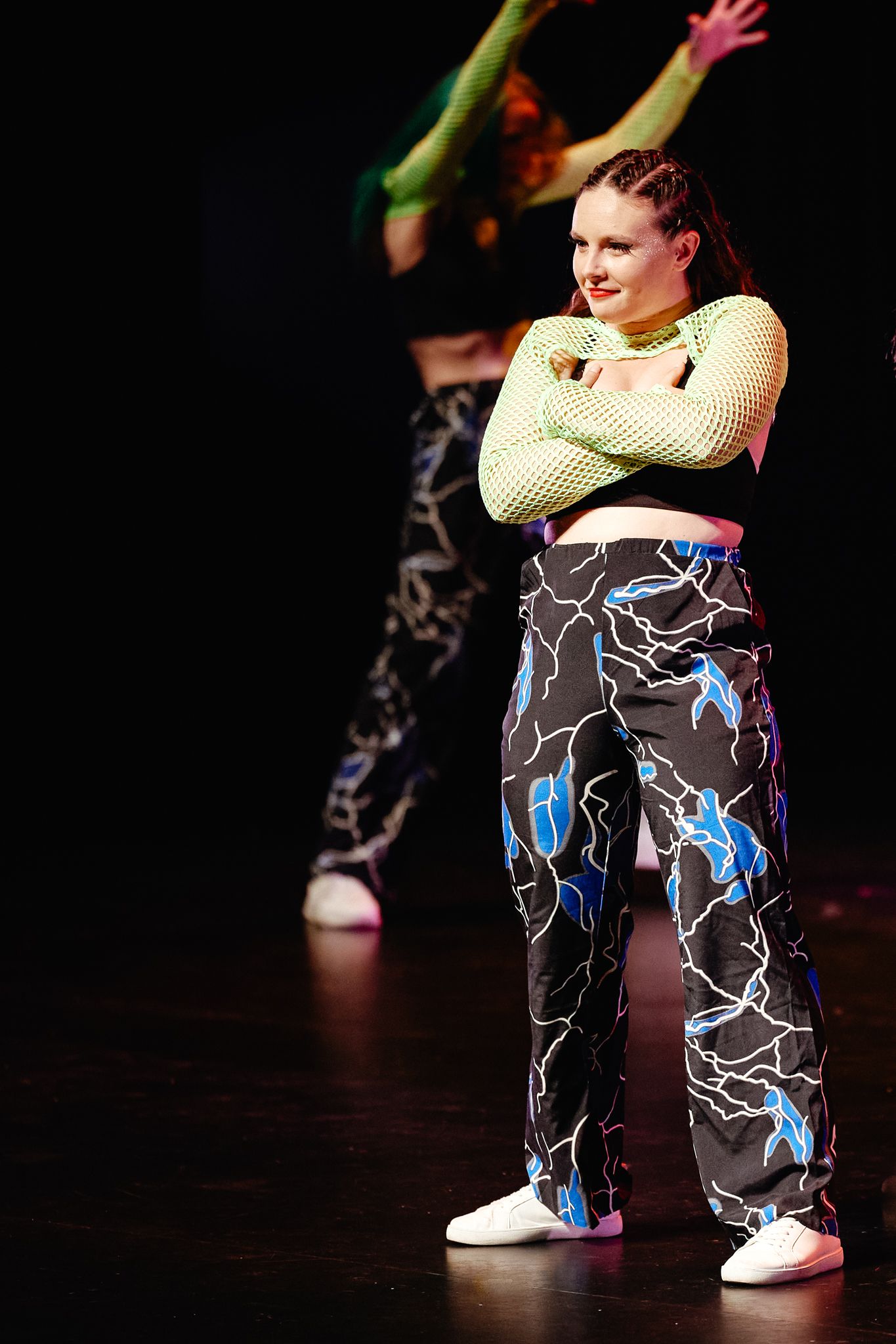 Performer in a mesh top and patterned pants strikes a dance pose on a dark stage during a performance.