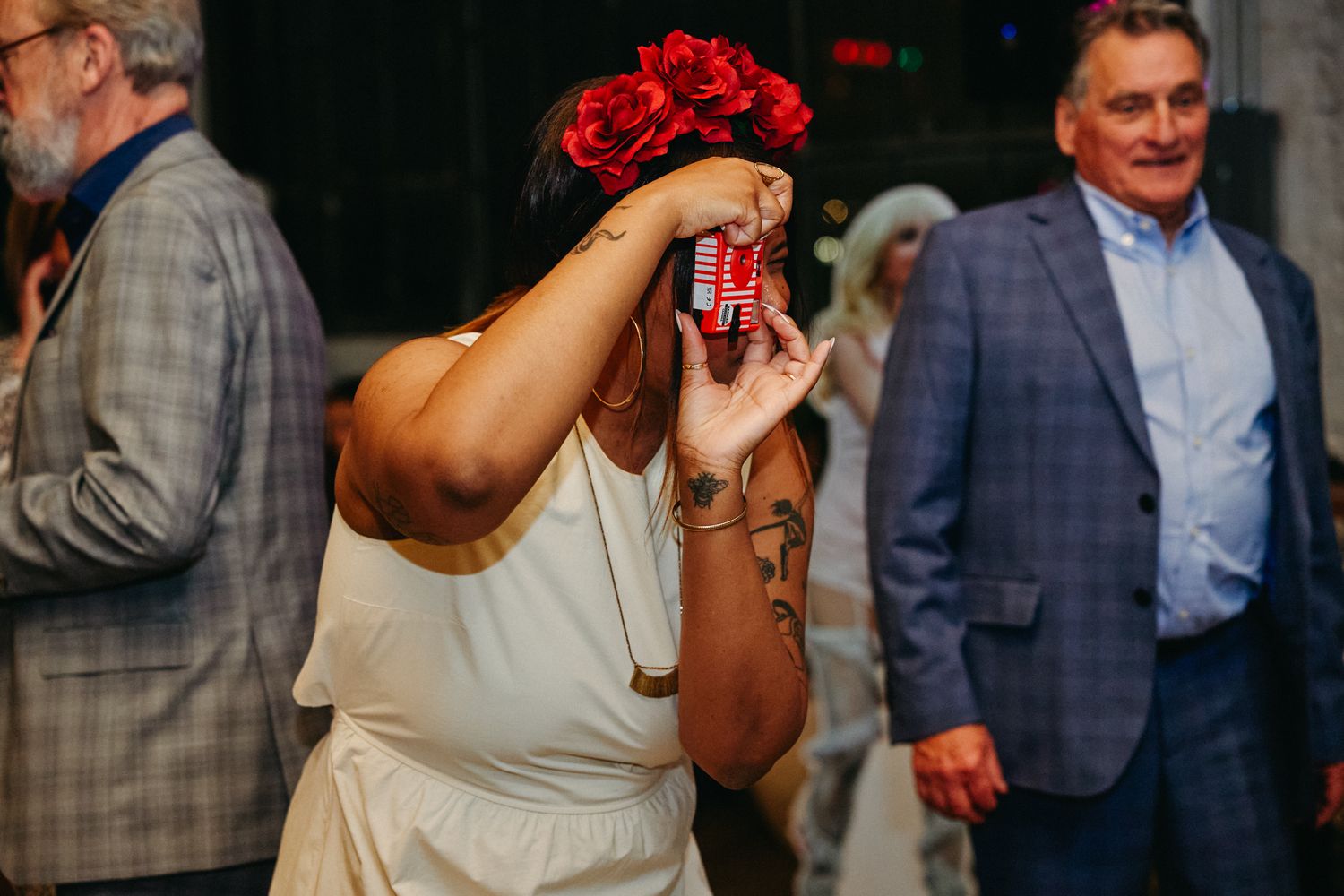 A wedding guest wearing a white dress and red flower crown takes a photo with a camera at a celebration event.