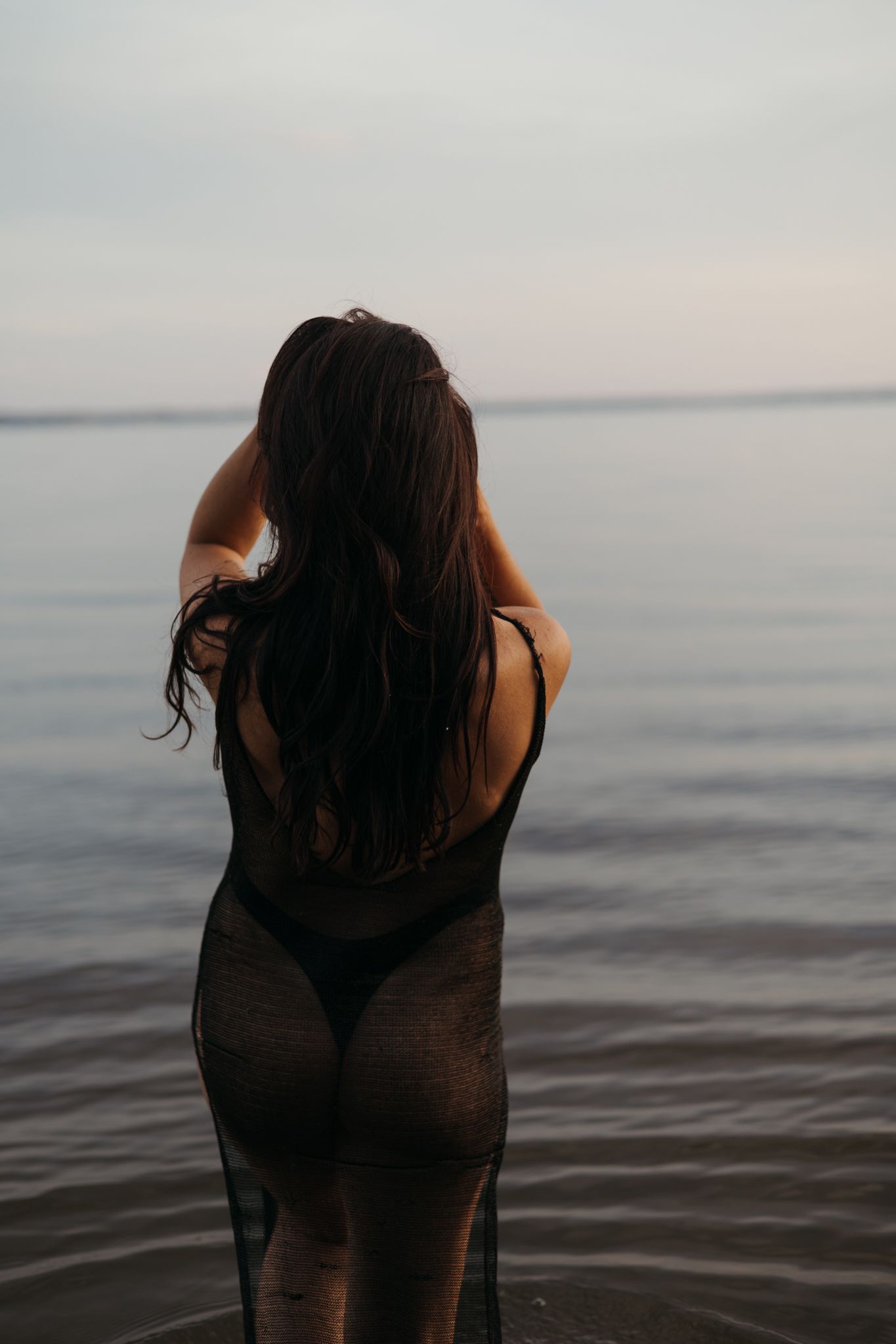 A silhouetted figure stands in calm water against an overcast sky at dusk.