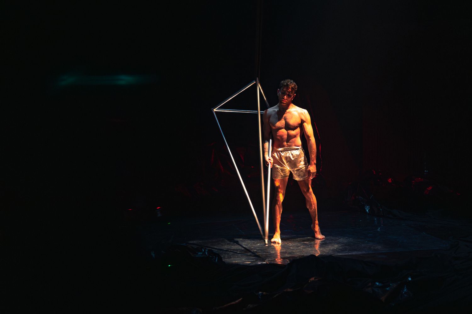 A performer stands in dramatic lighting next to a geometric light sculpture on a dark stage during an artistic performance.