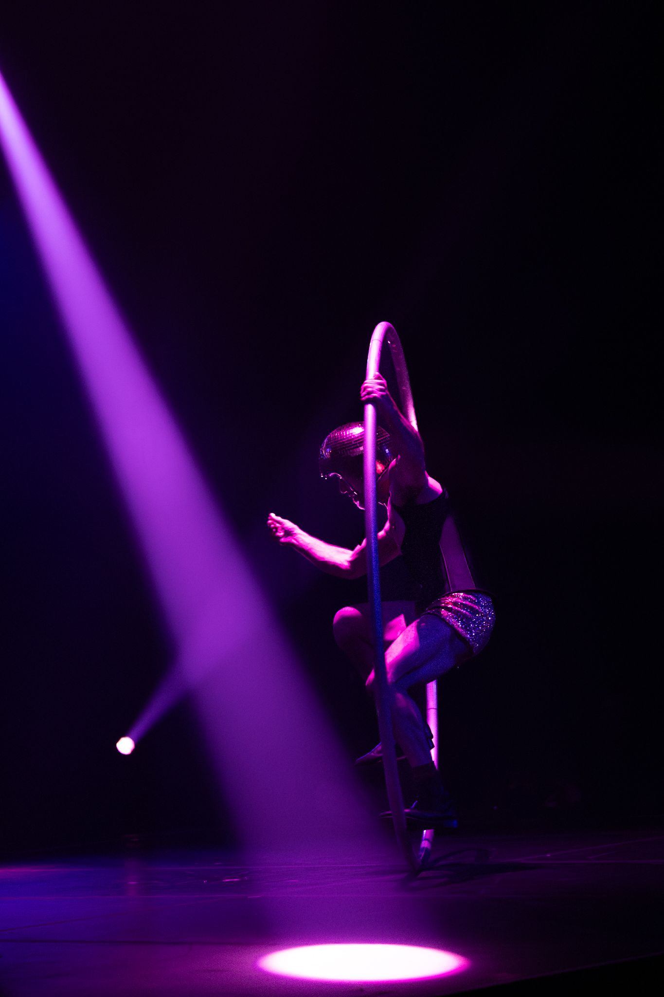 A pole dancer performs an artistic aerial move under dramatic purple stage lighting in a dark theater setting.