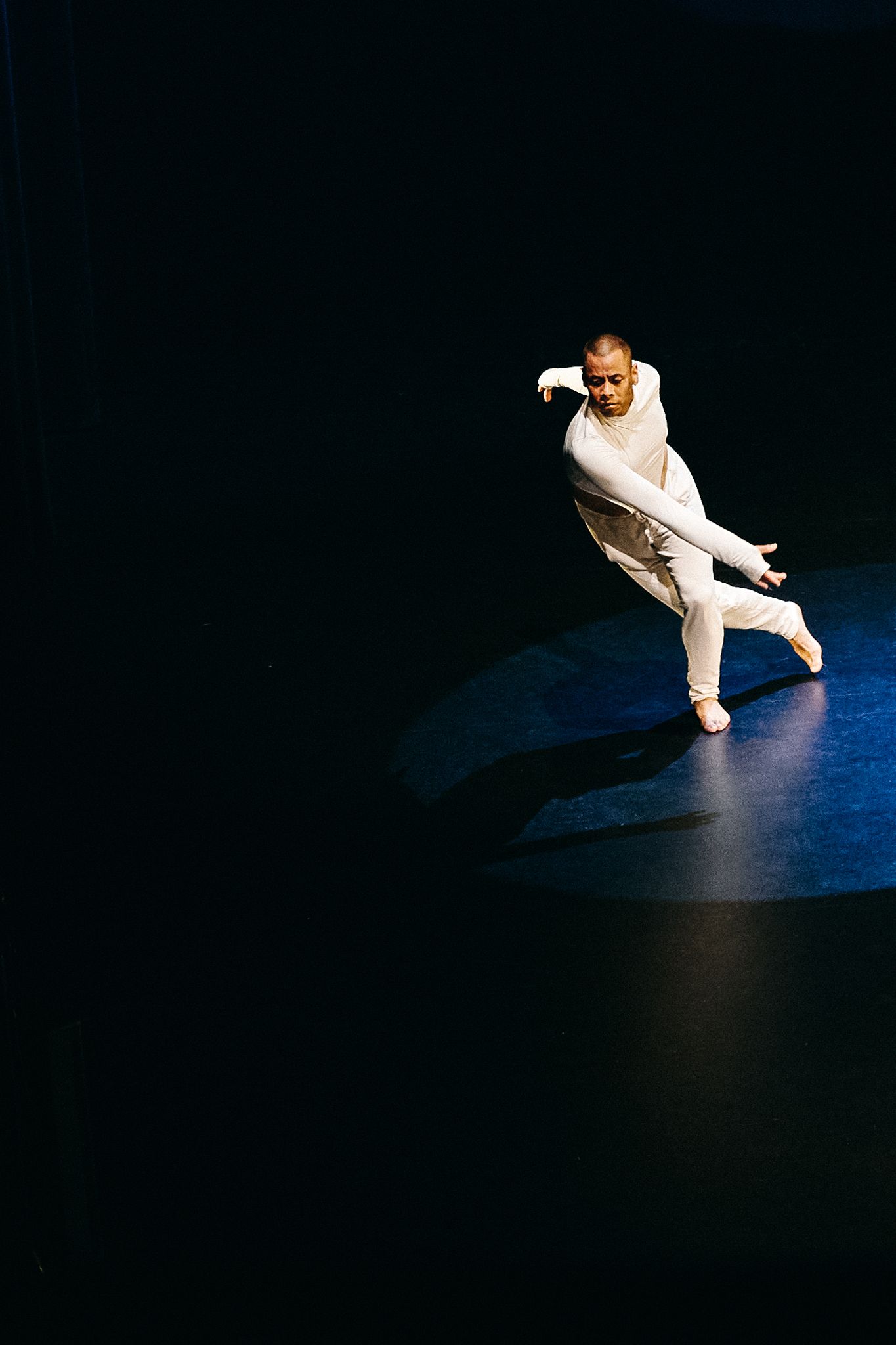 A dancer in white performs a low crouching movement sequence against a dark background in contemporary dance style.