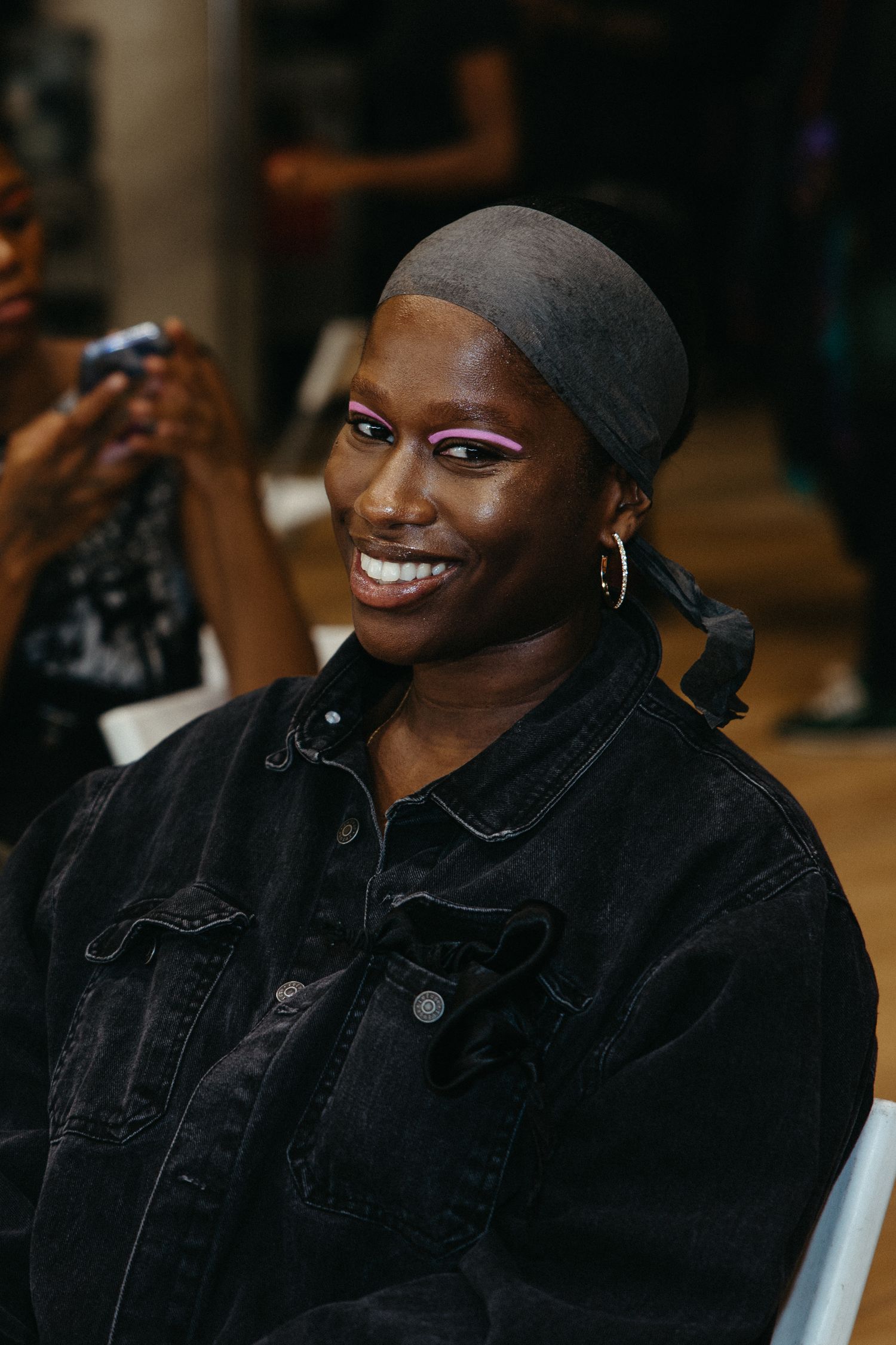 A person wearing a black denim jacket and head scarf smiles warmly at the camera.