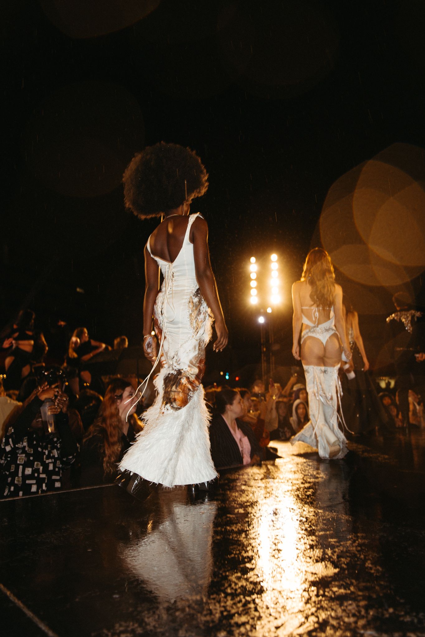 Models in white dresses walk a dimly lit runway at night as flames illuminate the scene.