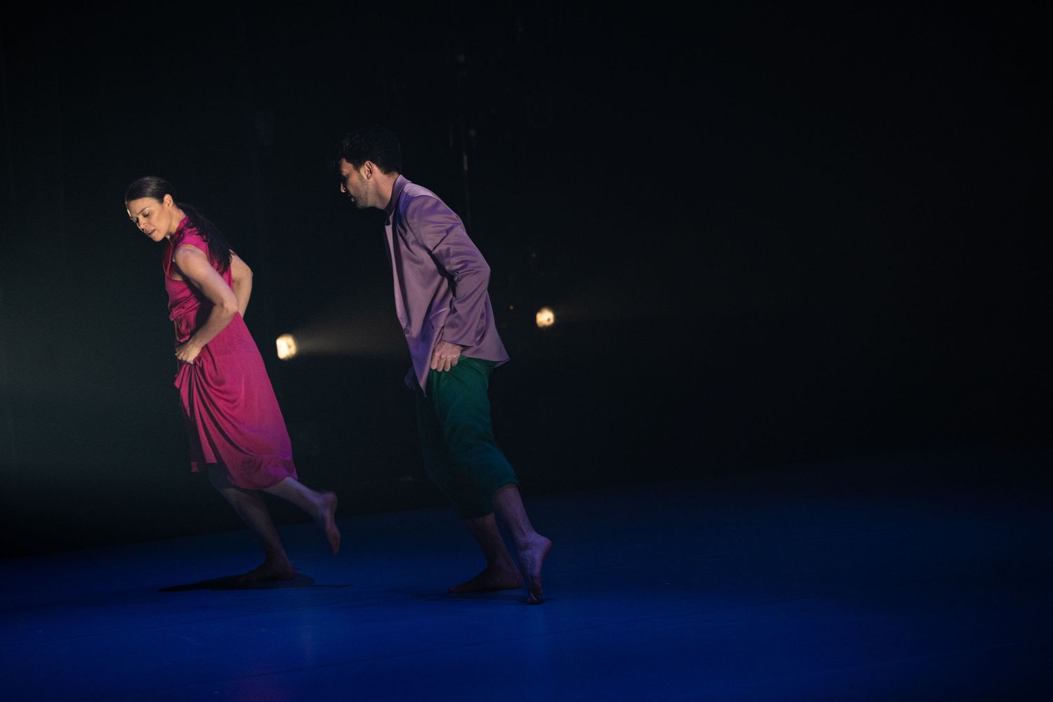 Two dancers in vibrant clothing perform an energetic synchronized dance routine on a dark stage.