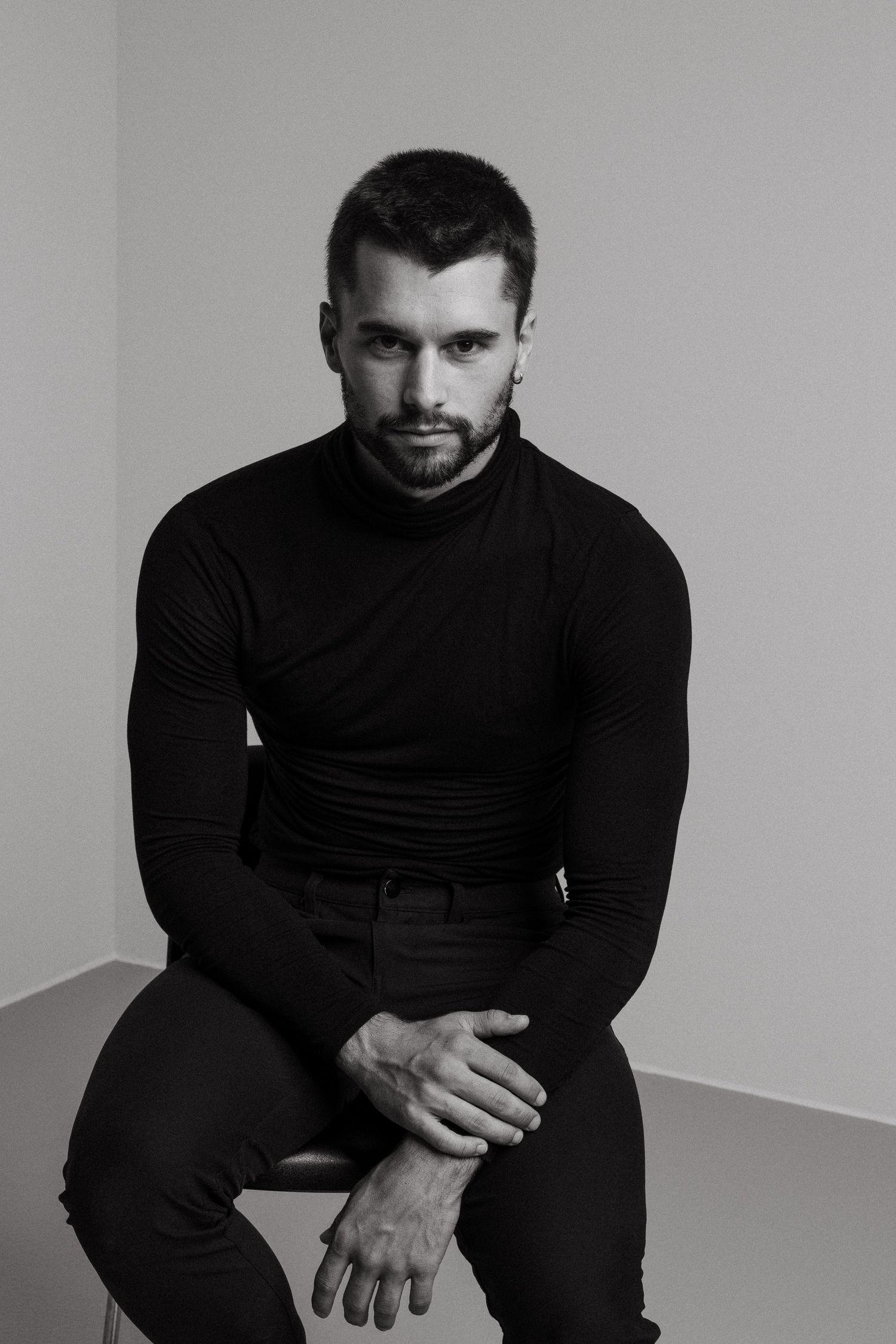 Black and white portrait of a person in dark turtleneck and pants sitting against a white corner wall.