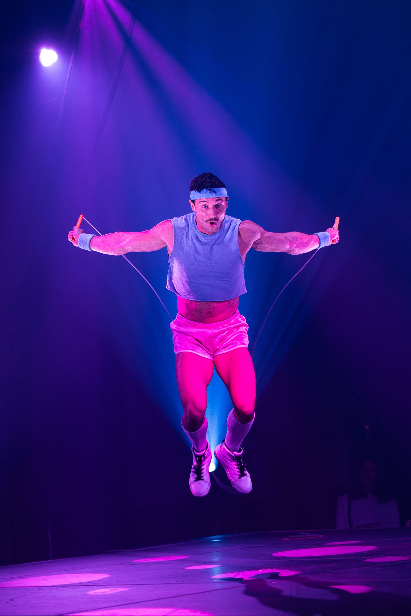 Performer in bright clothing leaps high into the air under striking blue and purple stage lighting.