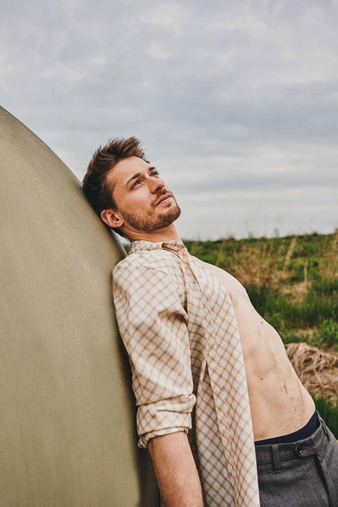 A person in a checkered shirt leans against a wall outdoors with dramatic cloudy sky and grassy field in background.