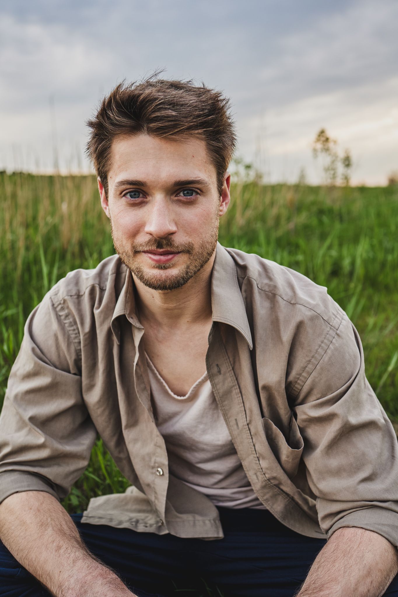 A person wearing a beige button-up shirt poses outdoors in a grassy field during an overcast day.