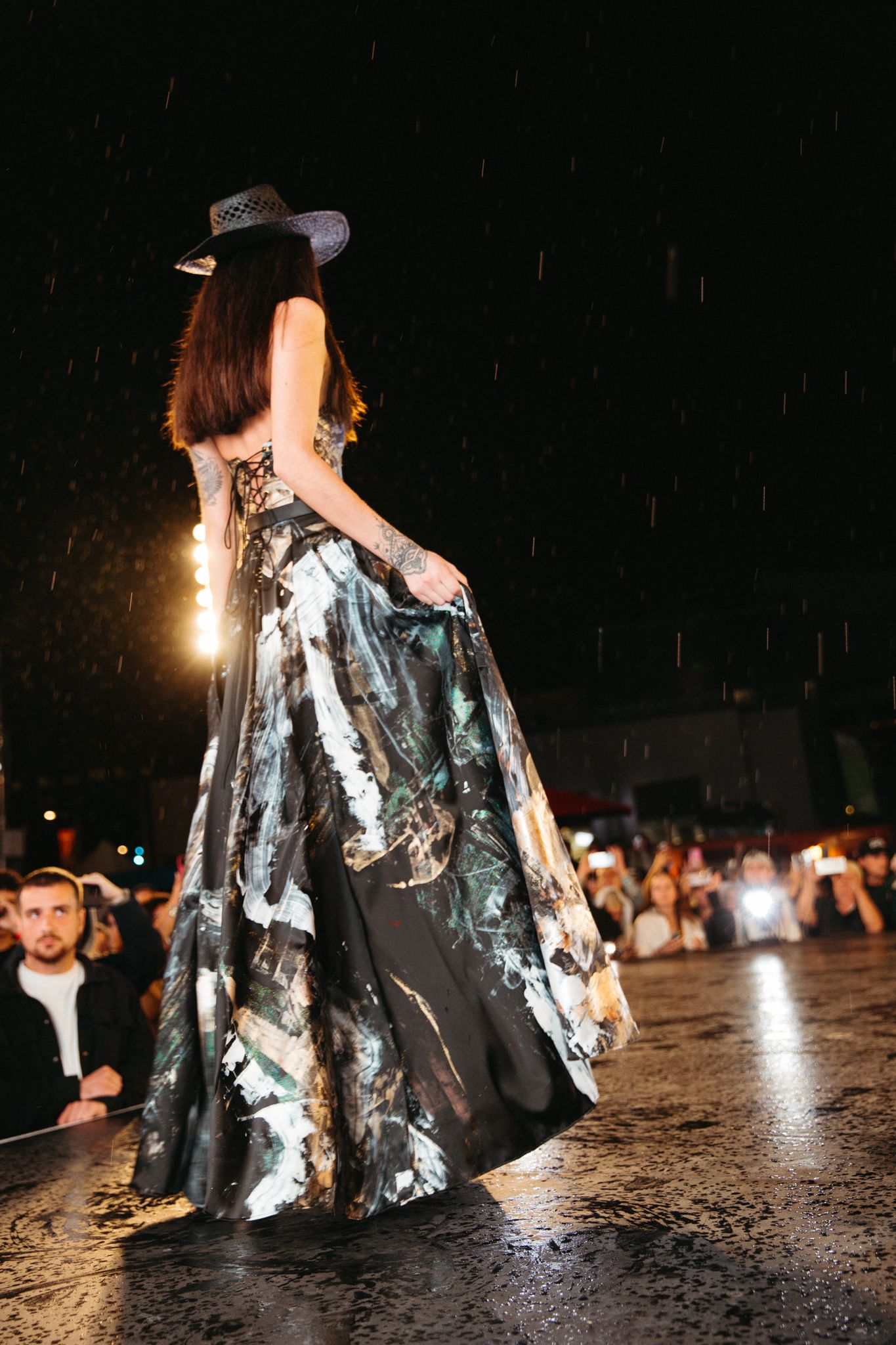 A fashion model walks down a dark runway wearing a dramatic silver and black evening gown with metallic accents.