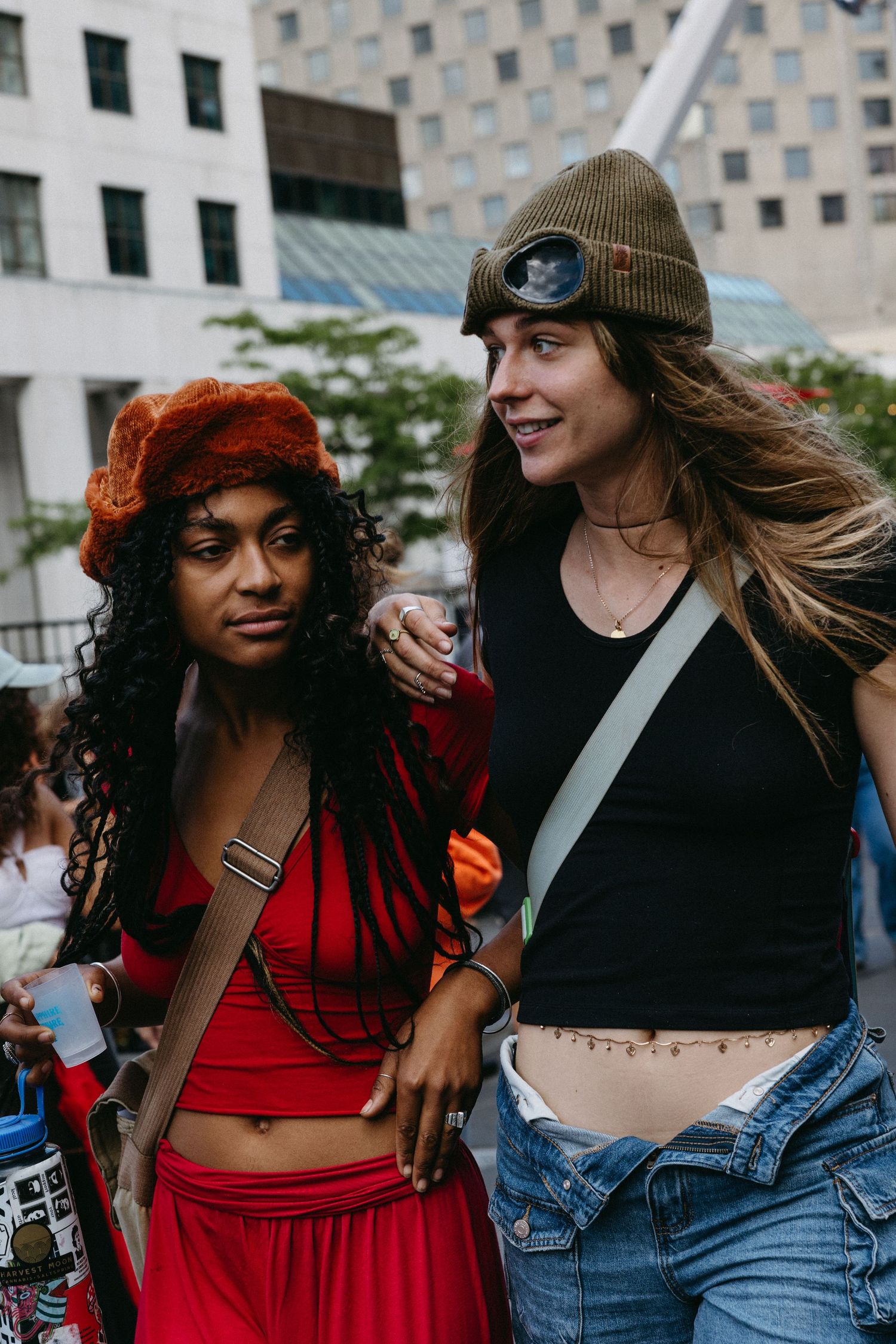 Two friends in casual urban wear smiling and embracing outdoors with a city building in the background.