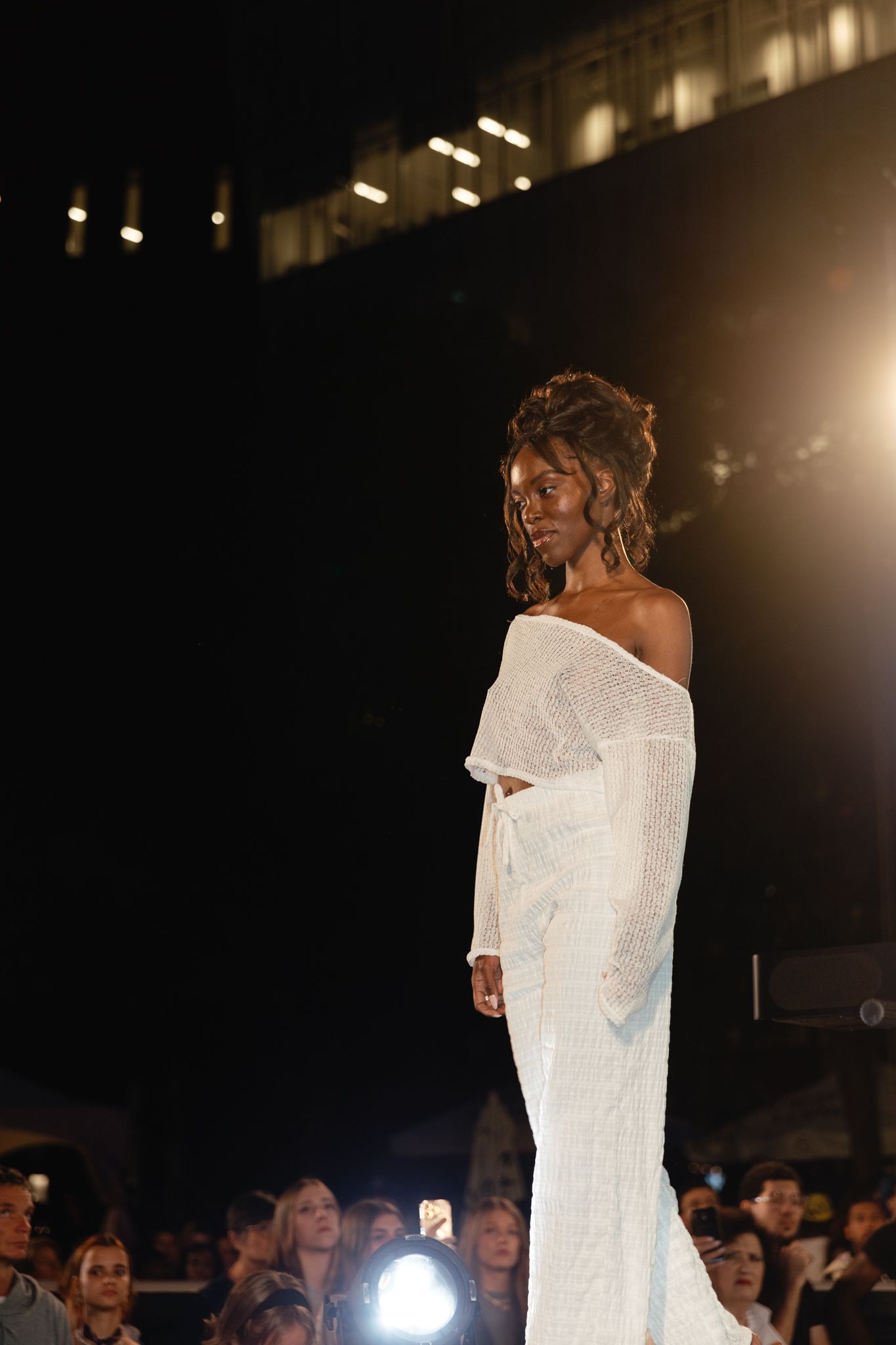 Fashion model walks runway in stunning off-shoulder white sequined evening gown at night fashion show.