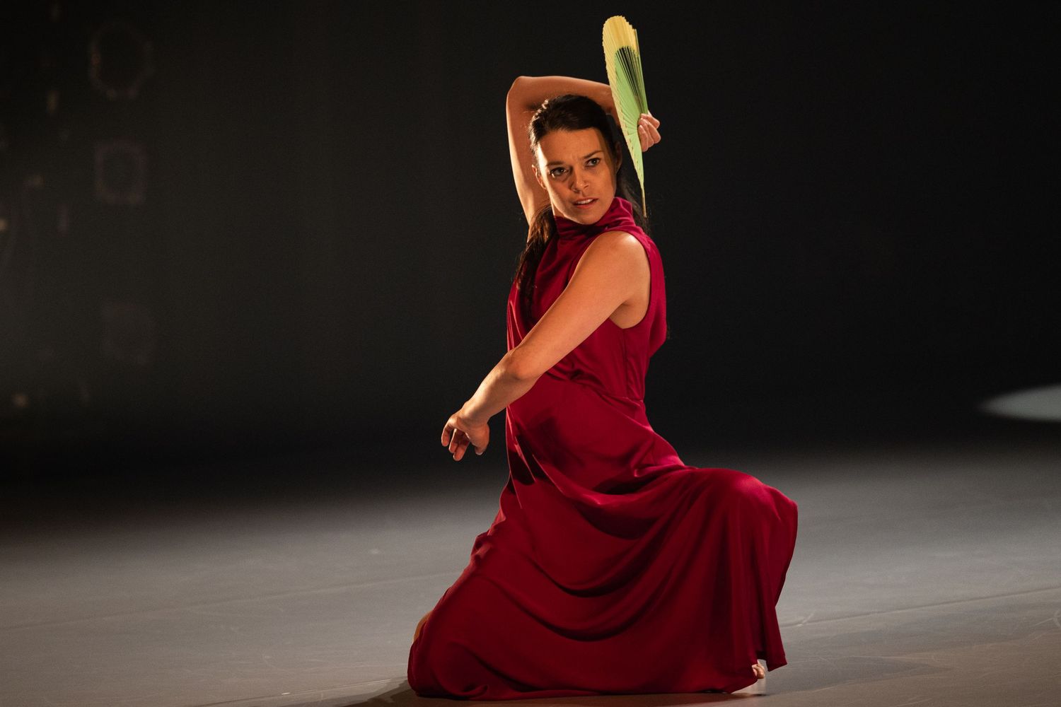Dancer in flowing red dress performs an expressive pose while holding yellow props against dark background.