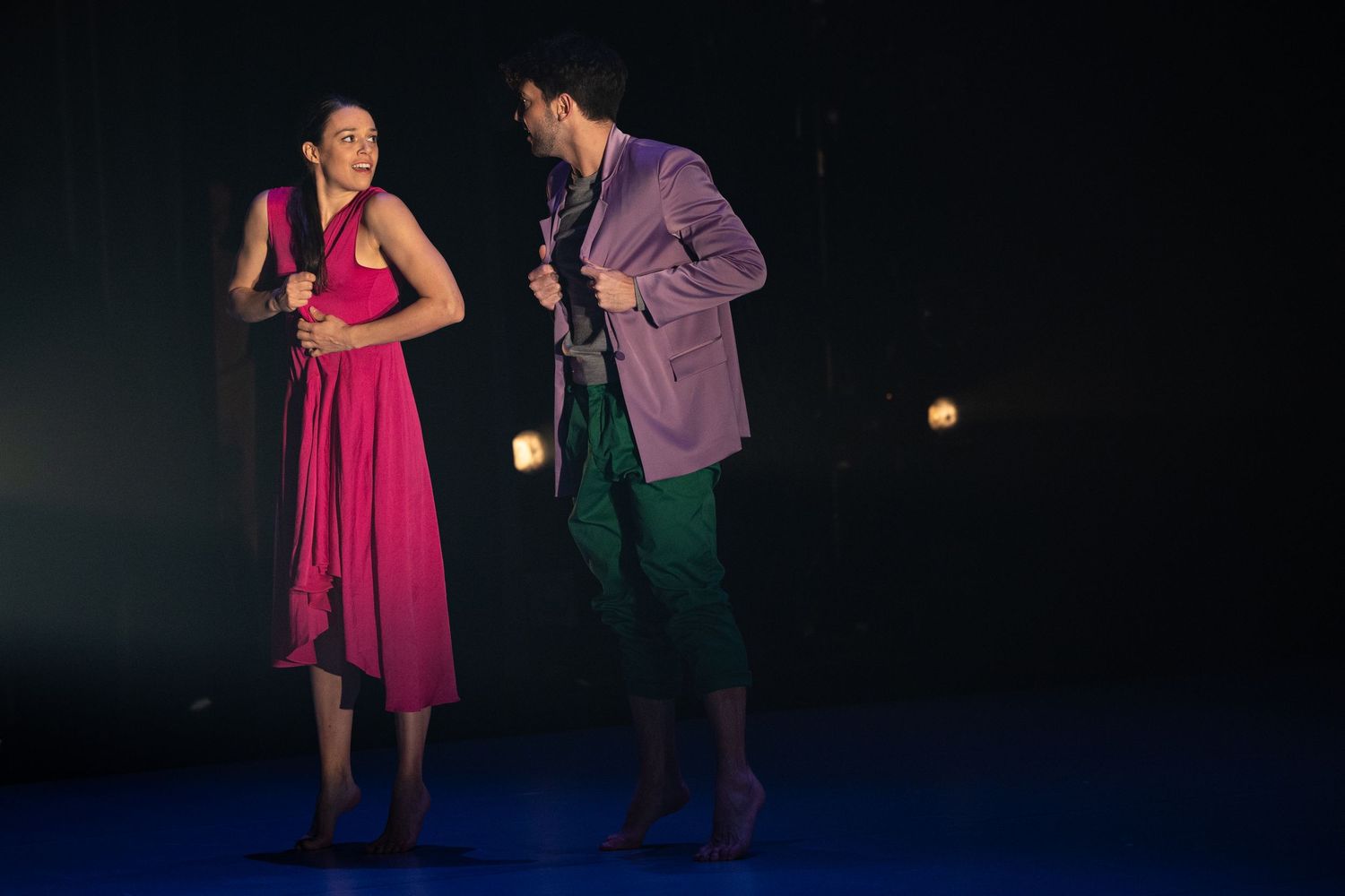 Performers in colorful dresses dance and engage in theatrical scenes during a stage performance lit by dramatic lighting.