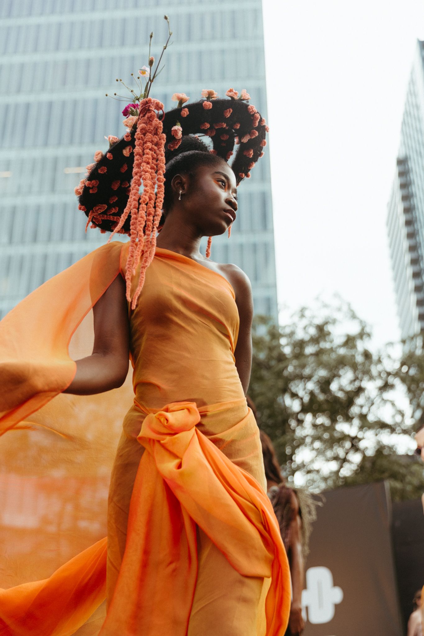 A figure in a flowing orange gown poses against urban architecture wearing statement floral headpieces.