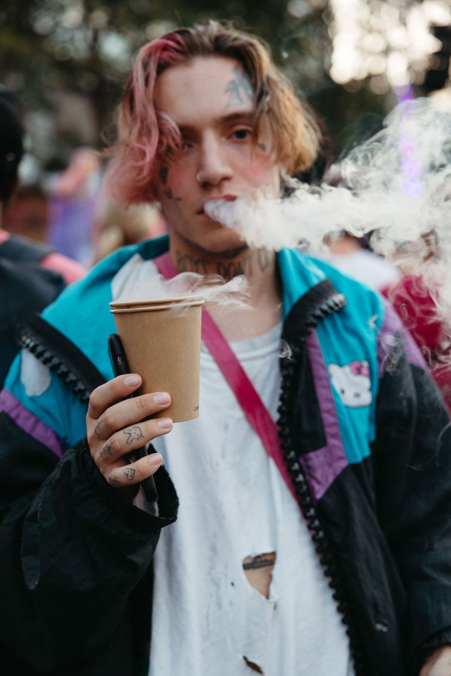 A person in a teal and purple jacket holds a coffee cup while exhaling vapor smoke in a casual outdoor setting.
