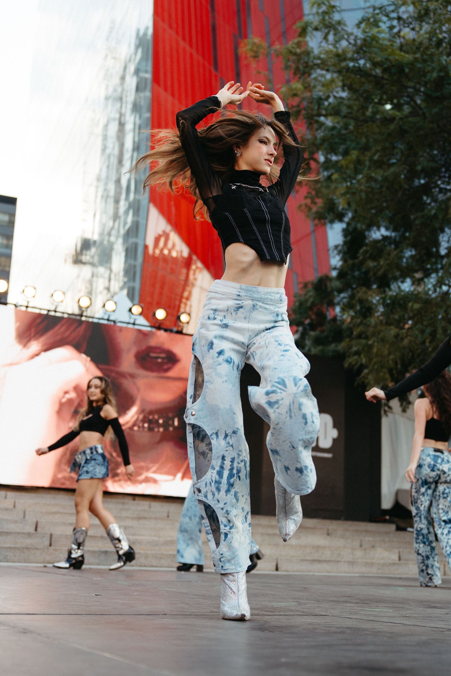Street dancer performing in ripped jeans and crop top near large red wall display with urban city backdrop.