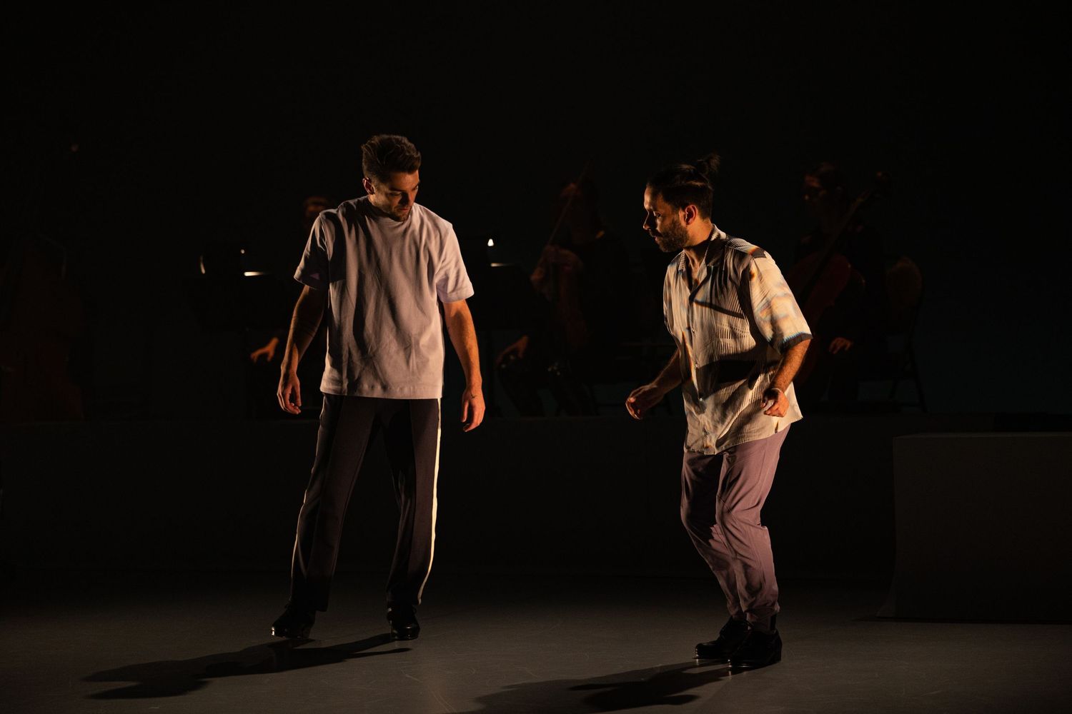 Two performers in casual attire crouch and dance during an expressive contemporary dance performance on a dark stage.