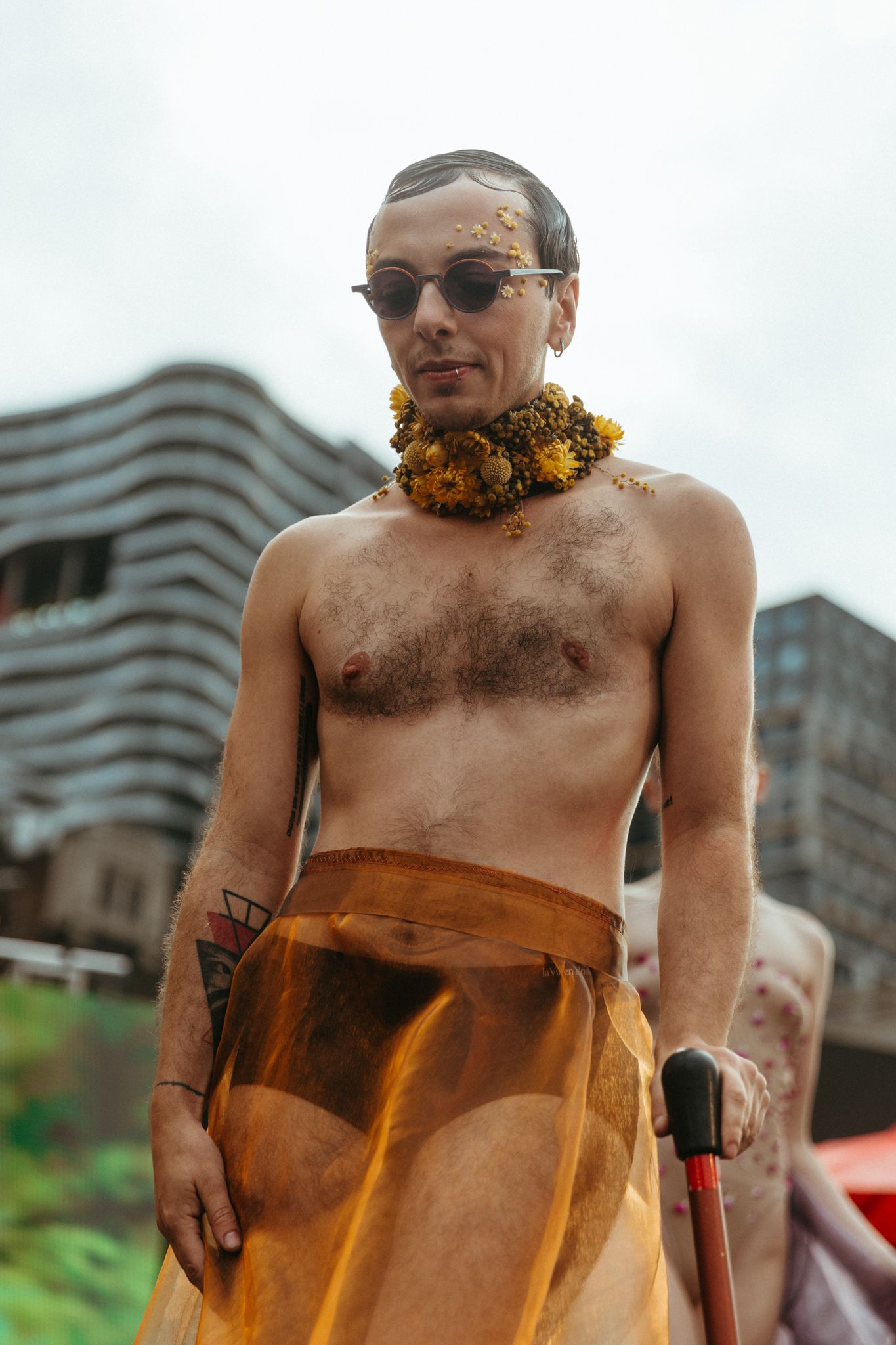 A shirtless person wearing sunglasses and a yellow flower crown stands outdoors near a modern building.