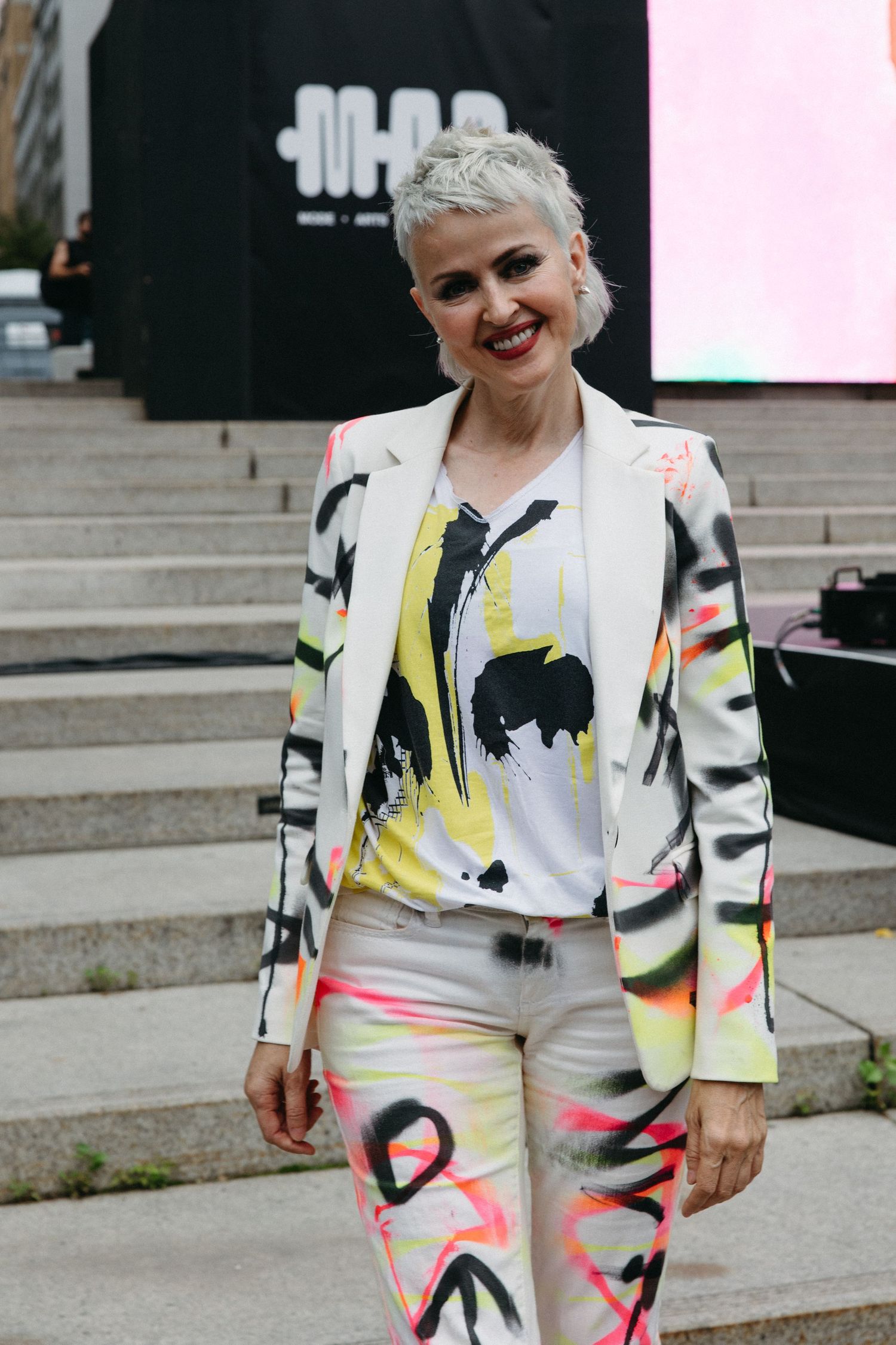 Bold artistic outfit featuring white suit with colorful graffiti-style paint splashes in yellow, black and pink on steps.