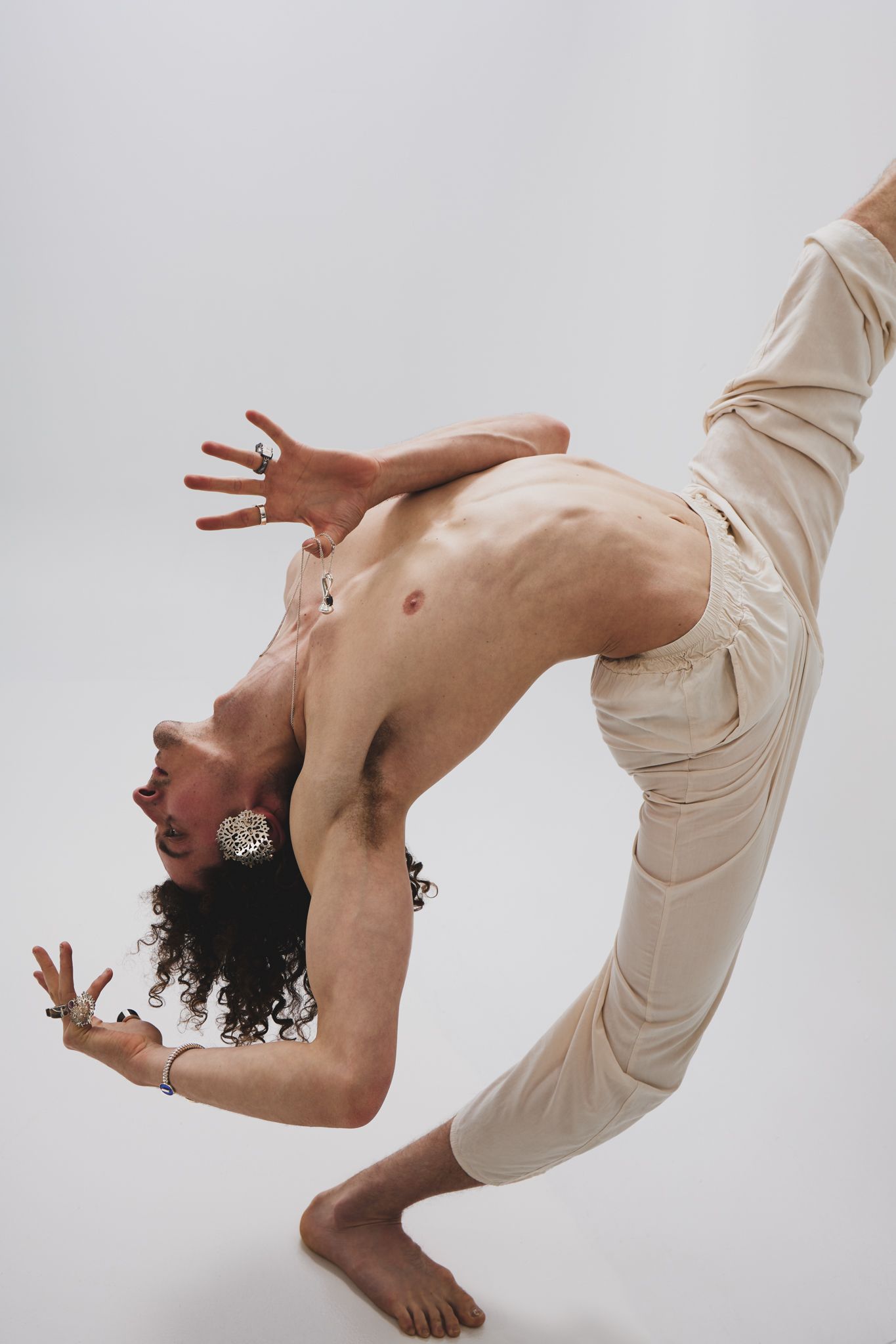 Contemporary dancer in cream colored clothing performs graceful backbend movements against white backdrop.