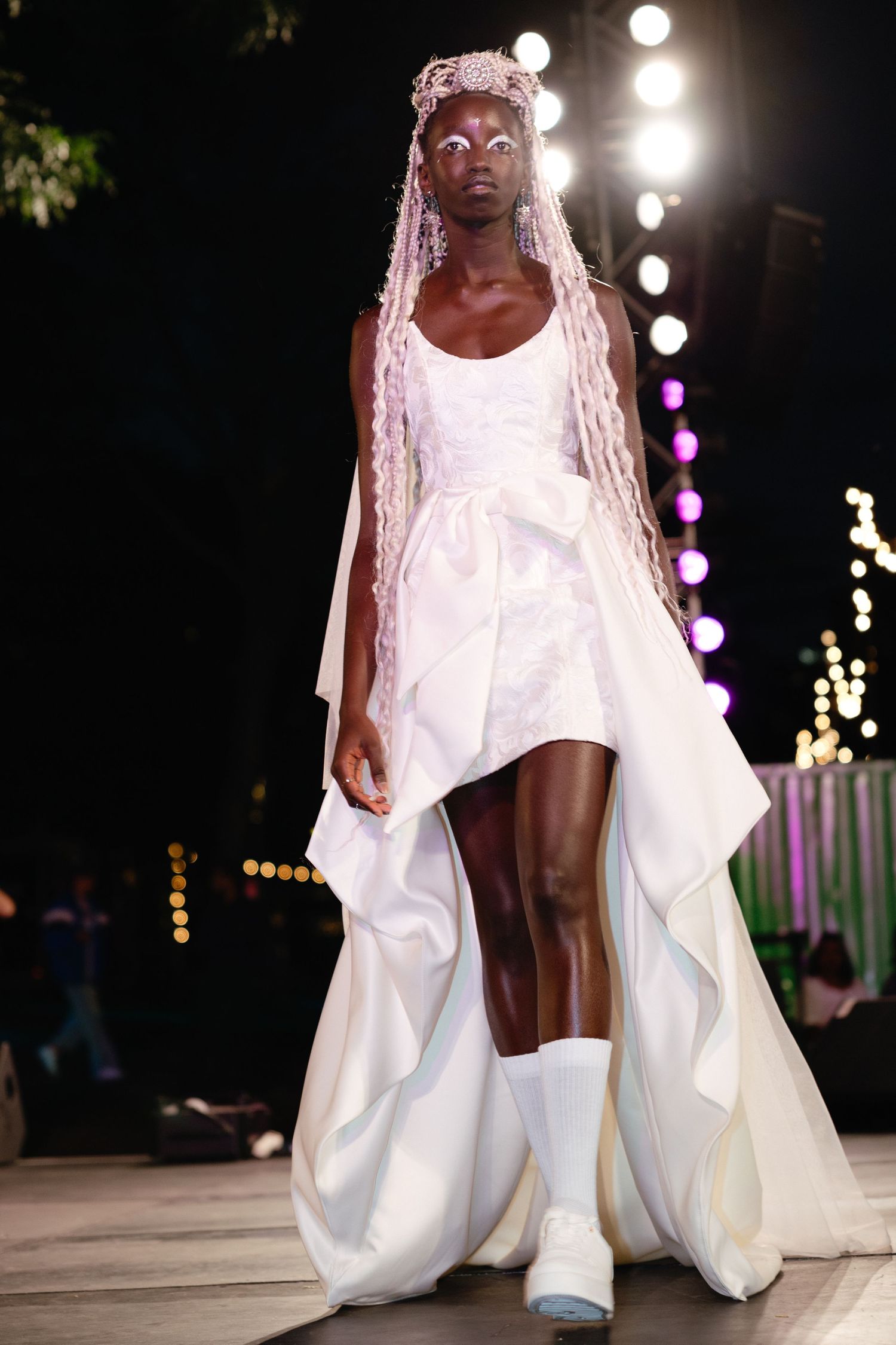 Model walks the runway in a flowing white wedding gown with dramatic fabric movement during a fashion show.