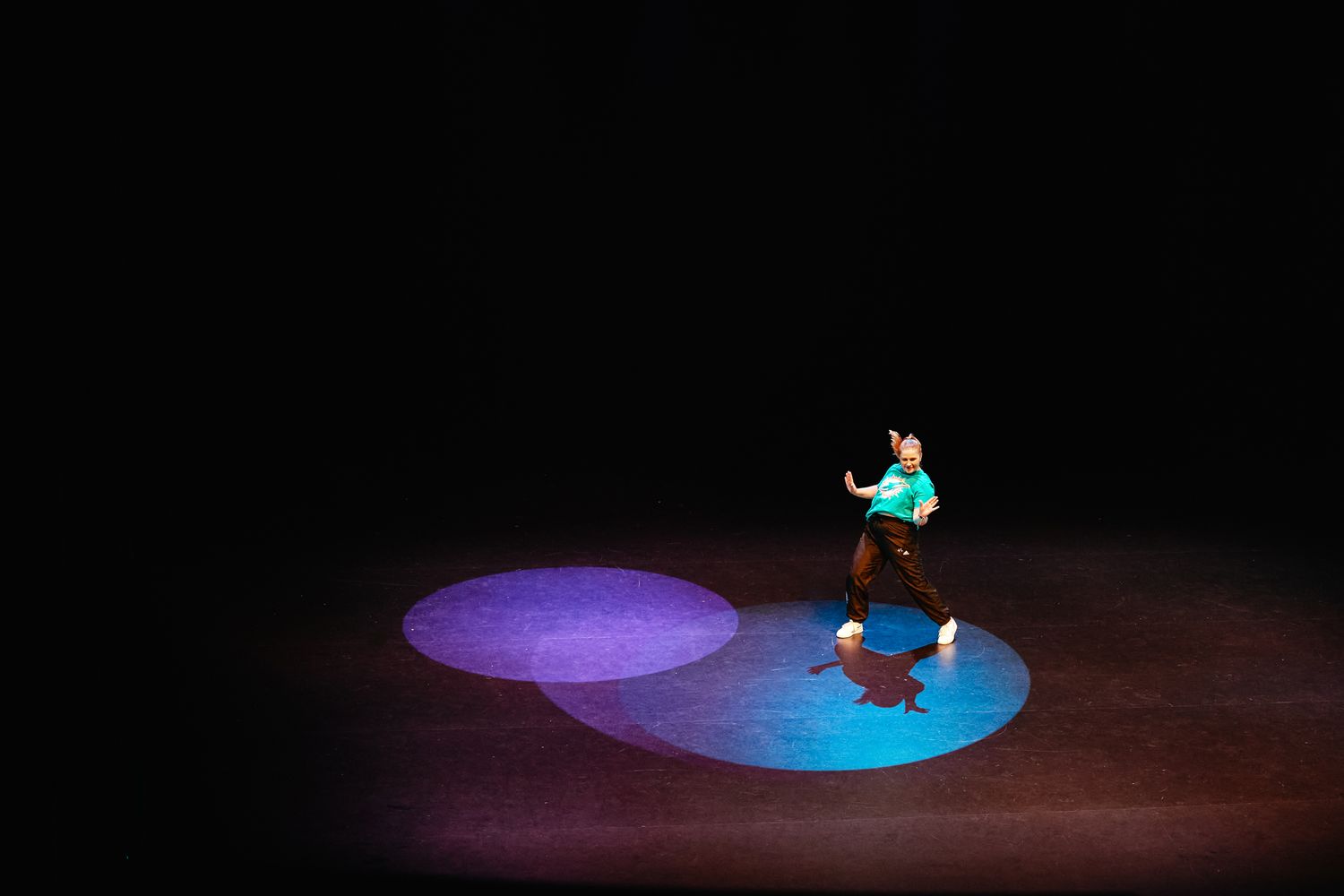A solo performer dances in a spotlight on a dark stage with purple and blue circular lighting effects on the floor.