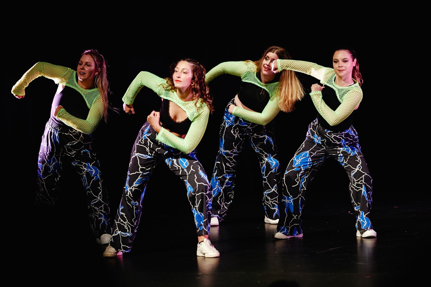 Dancers in blue and black costumes perform synchronized movements on a dark stage during a modern dance routine.