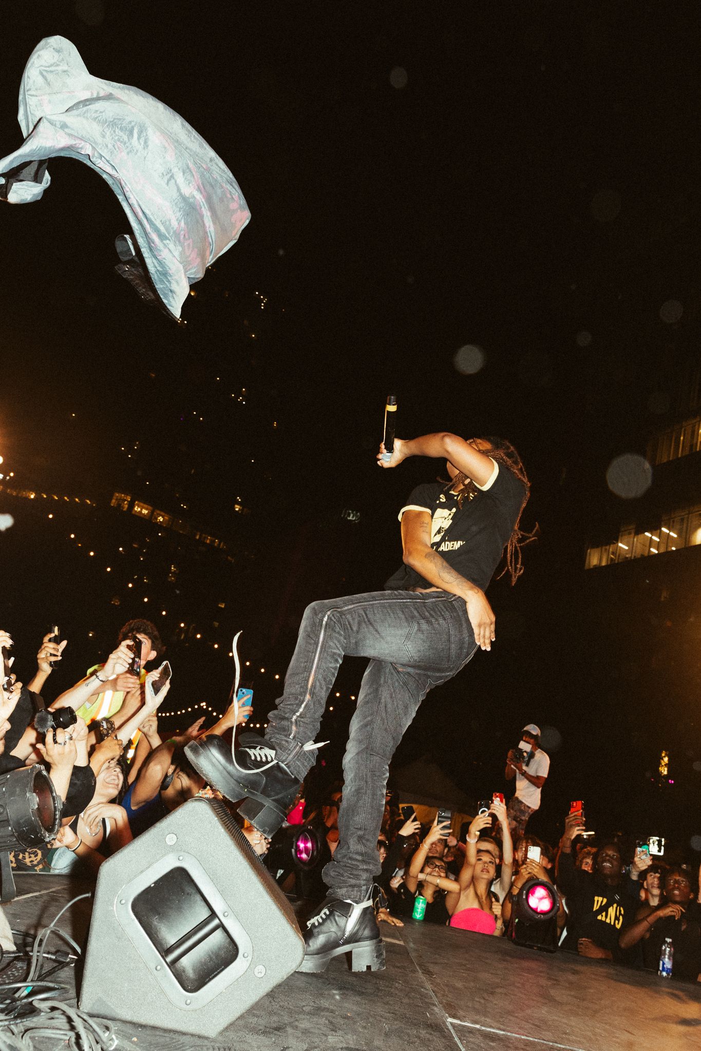 Performer jumps dramatically on stage with white fabric flying through the air as excited crowd watches at concert venue.