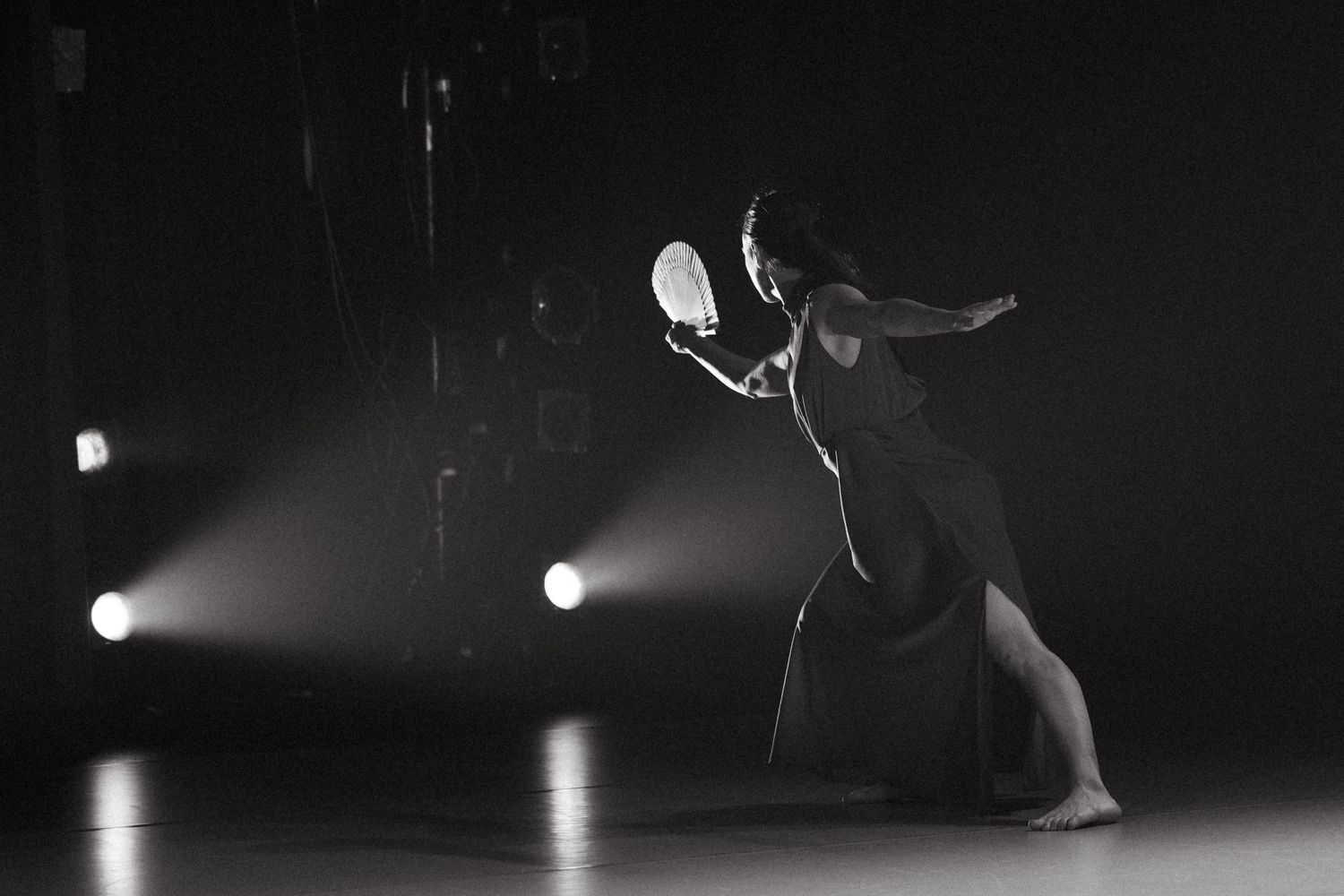Black and white performance photograph showing a solo dancer in motion on a dark stage under dramatic spotlights.
