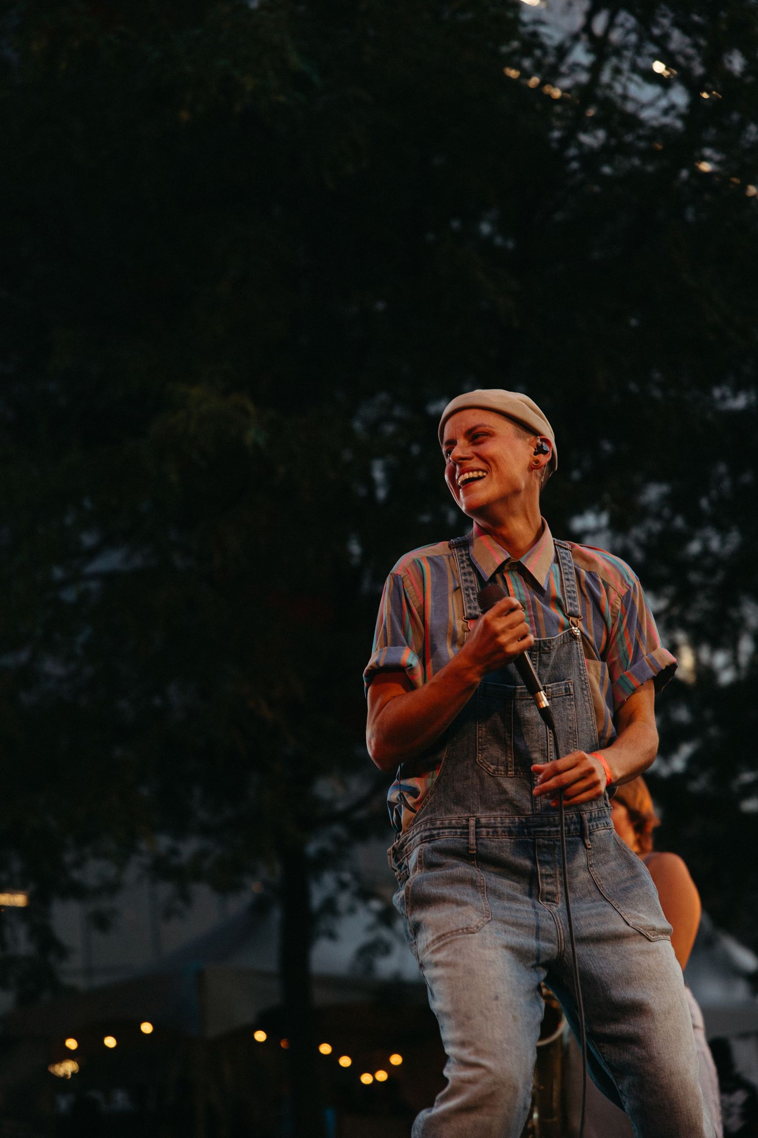 Performer in denim overalls and cap stands on stage at outdoor evening concert with twinkle lights in background.