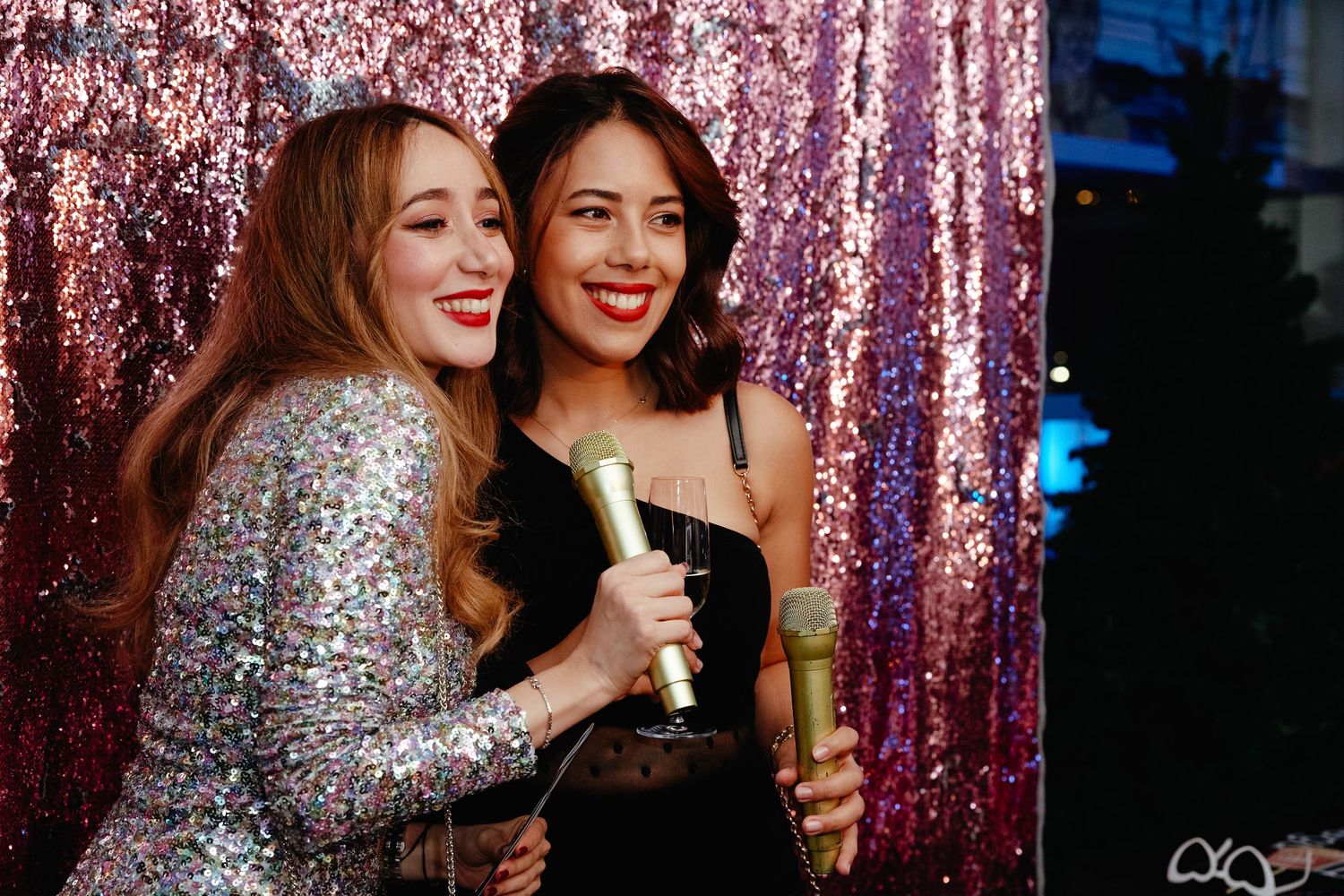 Two friends laugh together while posing with drinks against a sparkly pink sequin backdrop at a party.