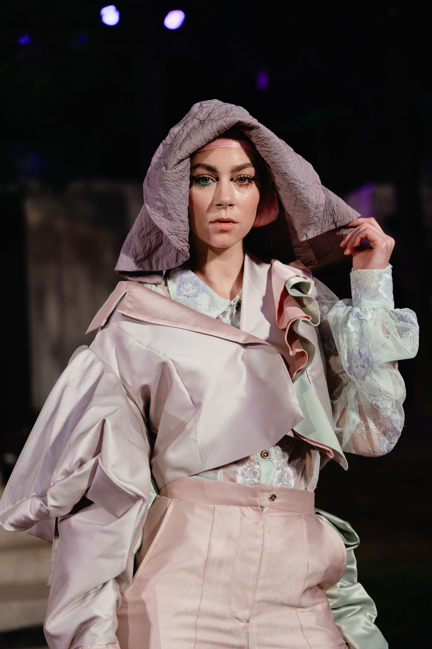 Model in light pink hooded ensemble with ruffled details poses on runway under stage lighting.