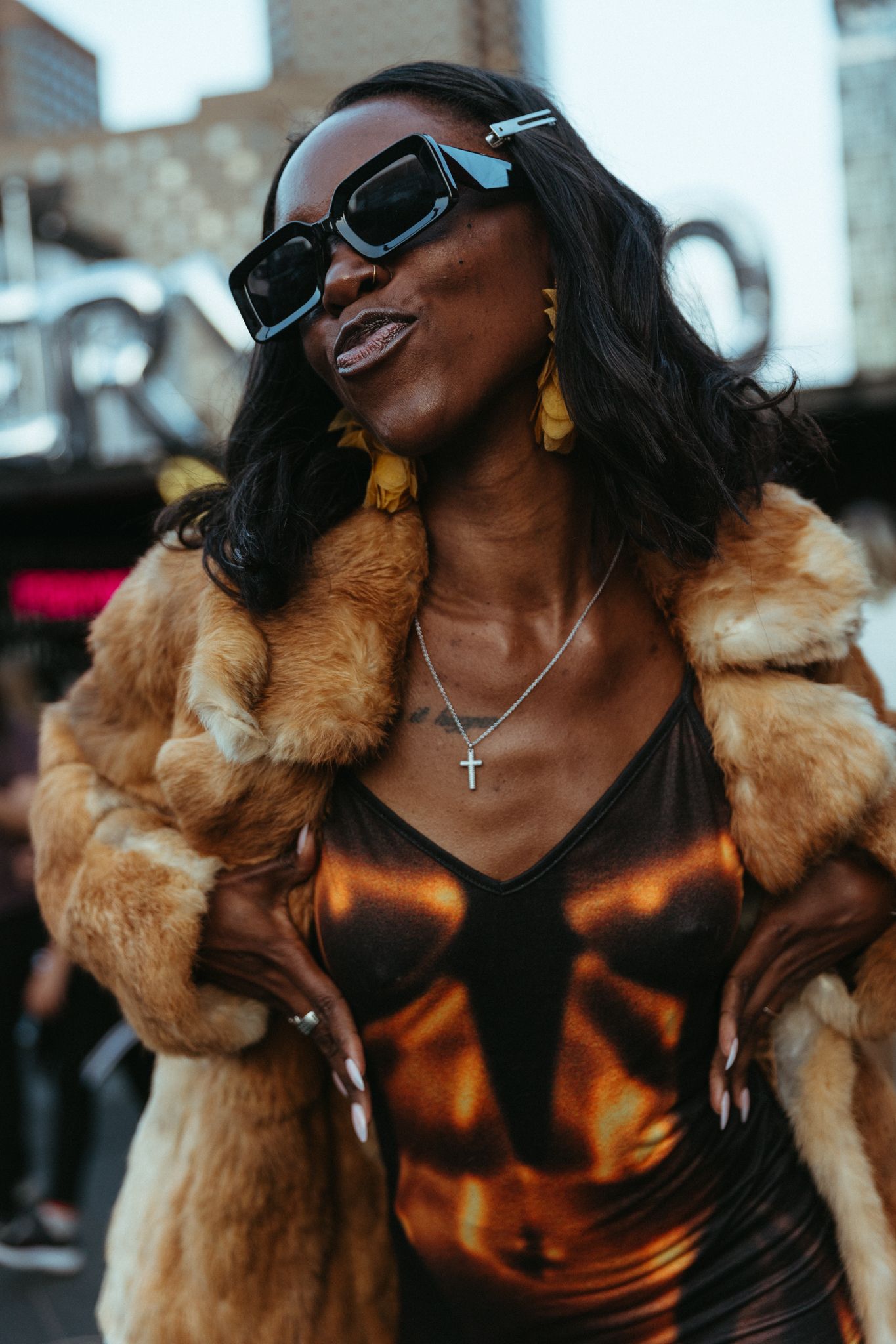 Stylish streetwear look featuring brown fur coat, metallic top, sunglasses and gold accessories in urban setting.