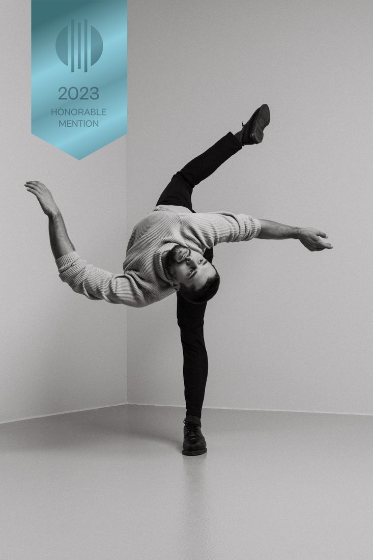 Dancer in casual attire performs graceful arabesque pose with outstretched limbs against minimalist studio backdrop.