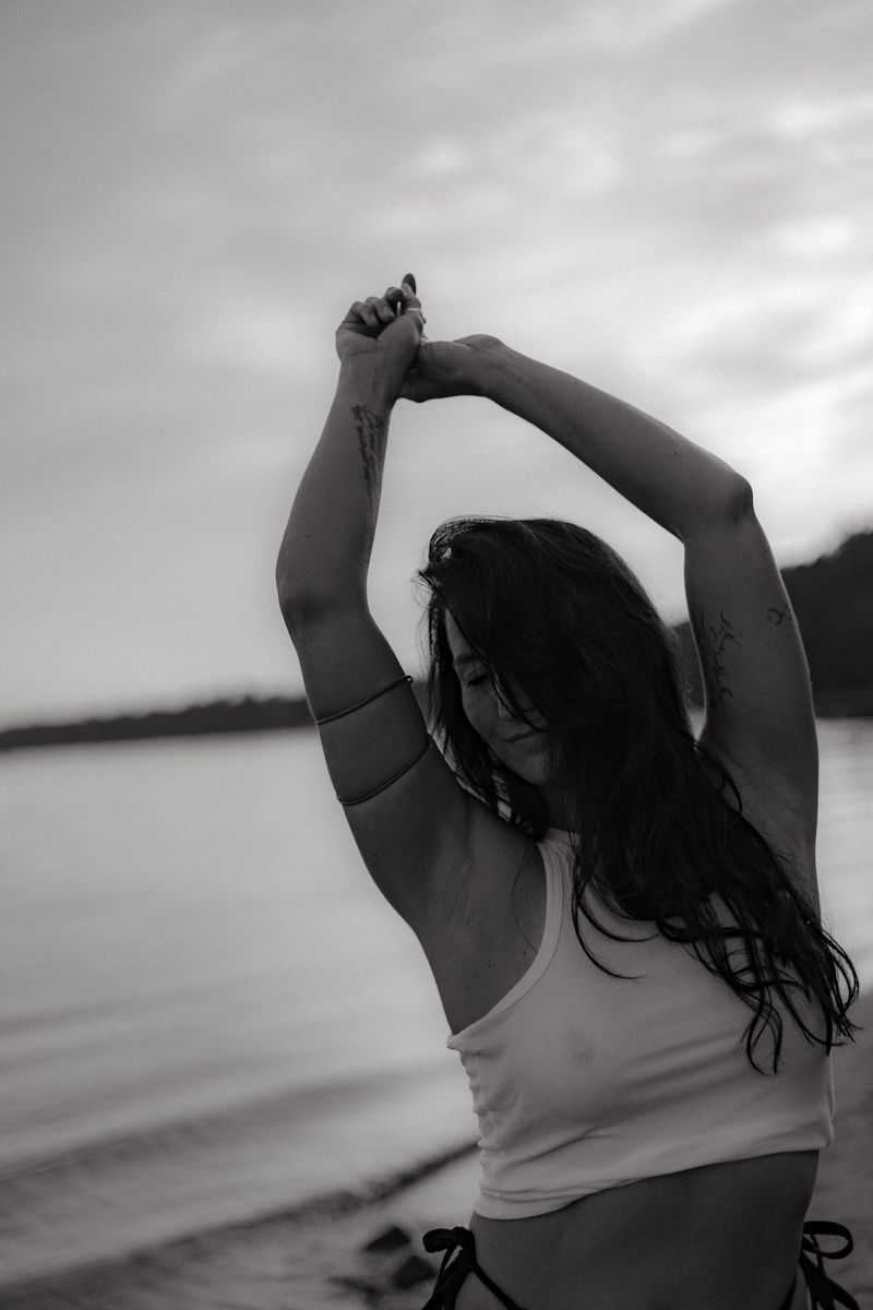 A silhouette of someone with long dark hair stretches their arms skyward at sunset by a peaceful lake.