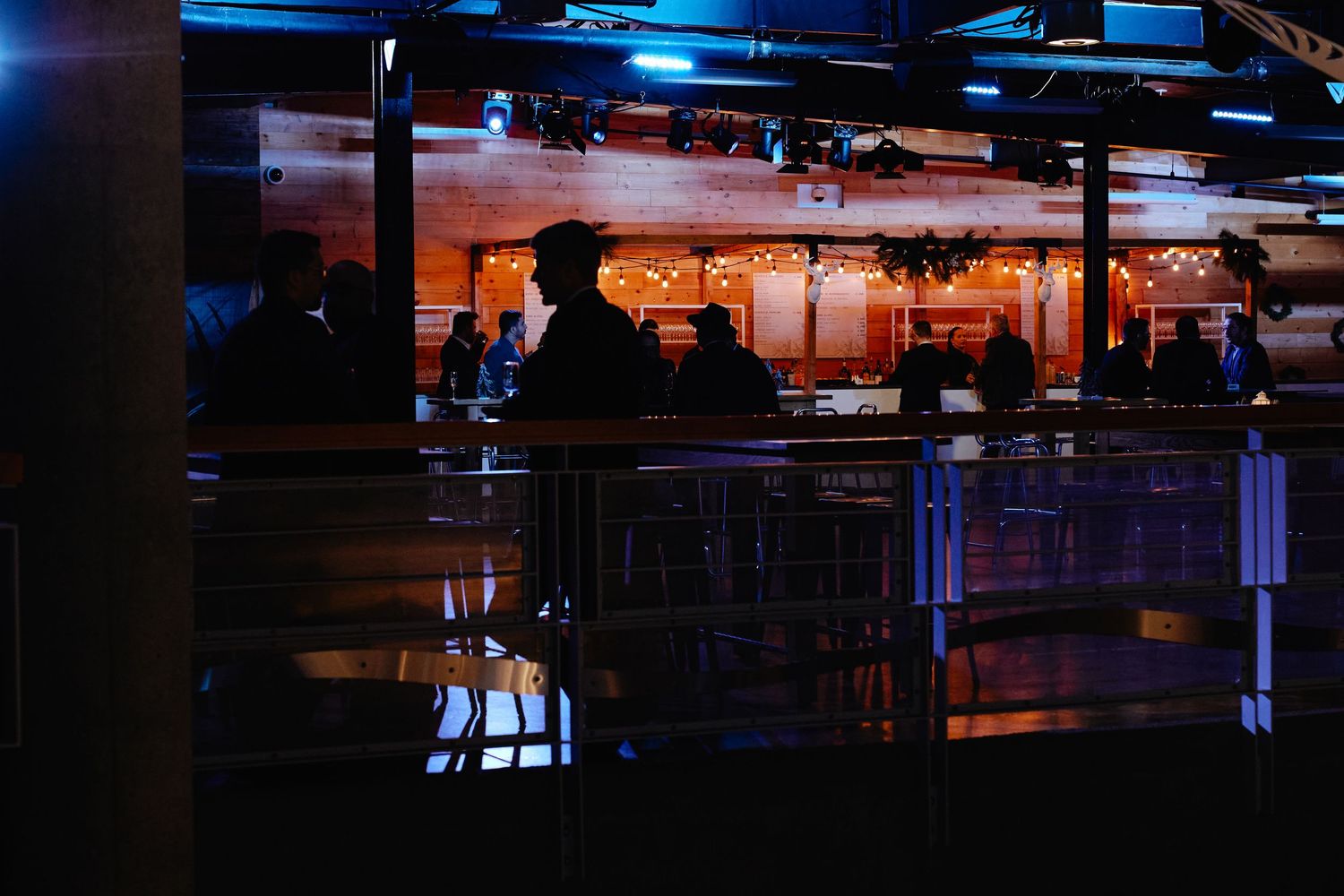 Dimly lit bar interior with blue ambient lighting and silhouettes reflected on glossy surfaces at night.