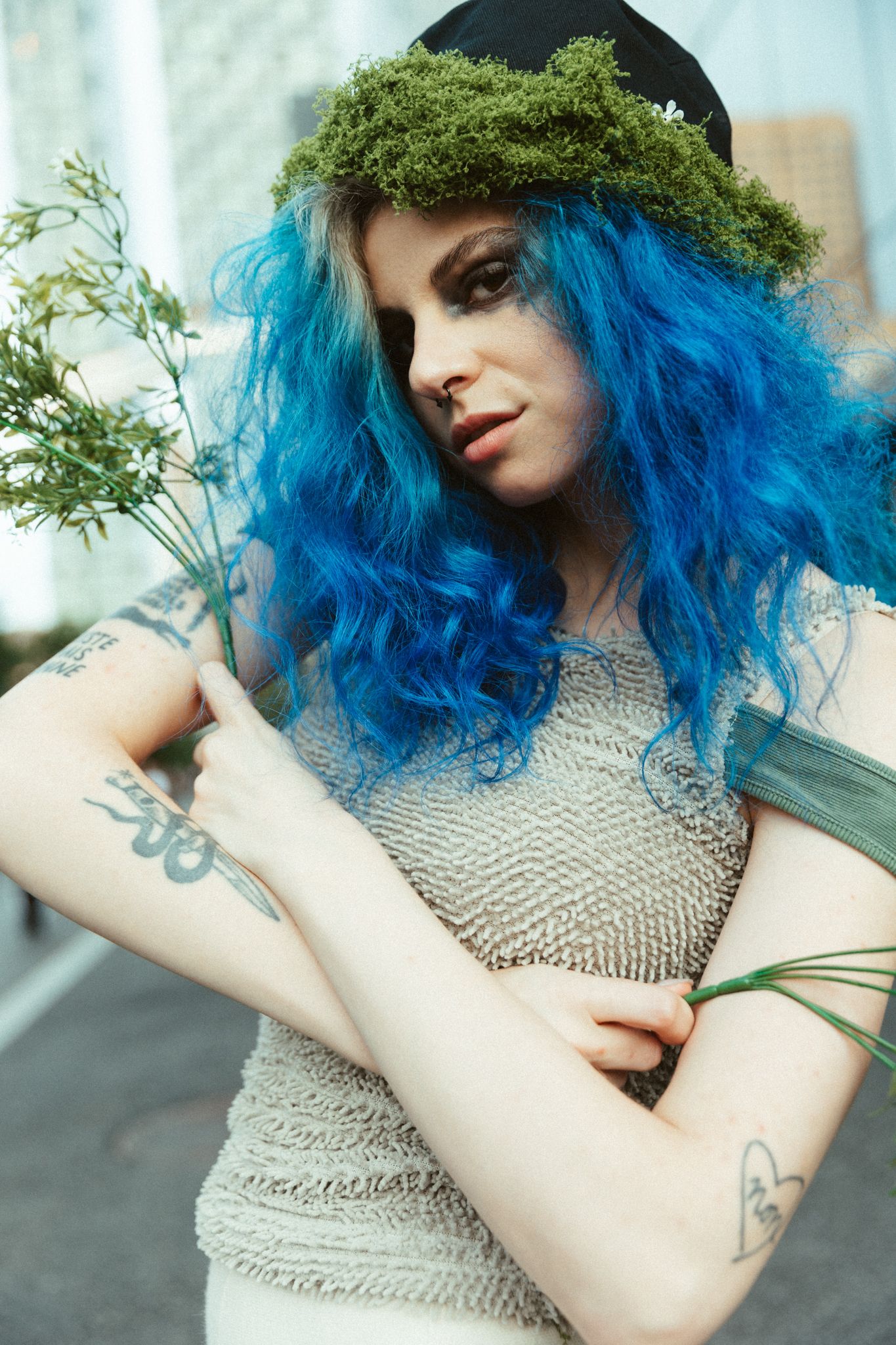 A fashion portrait features vibrant blue curly hair adorned with green moss, wearing a silvery mesh top and visible tattoos.
