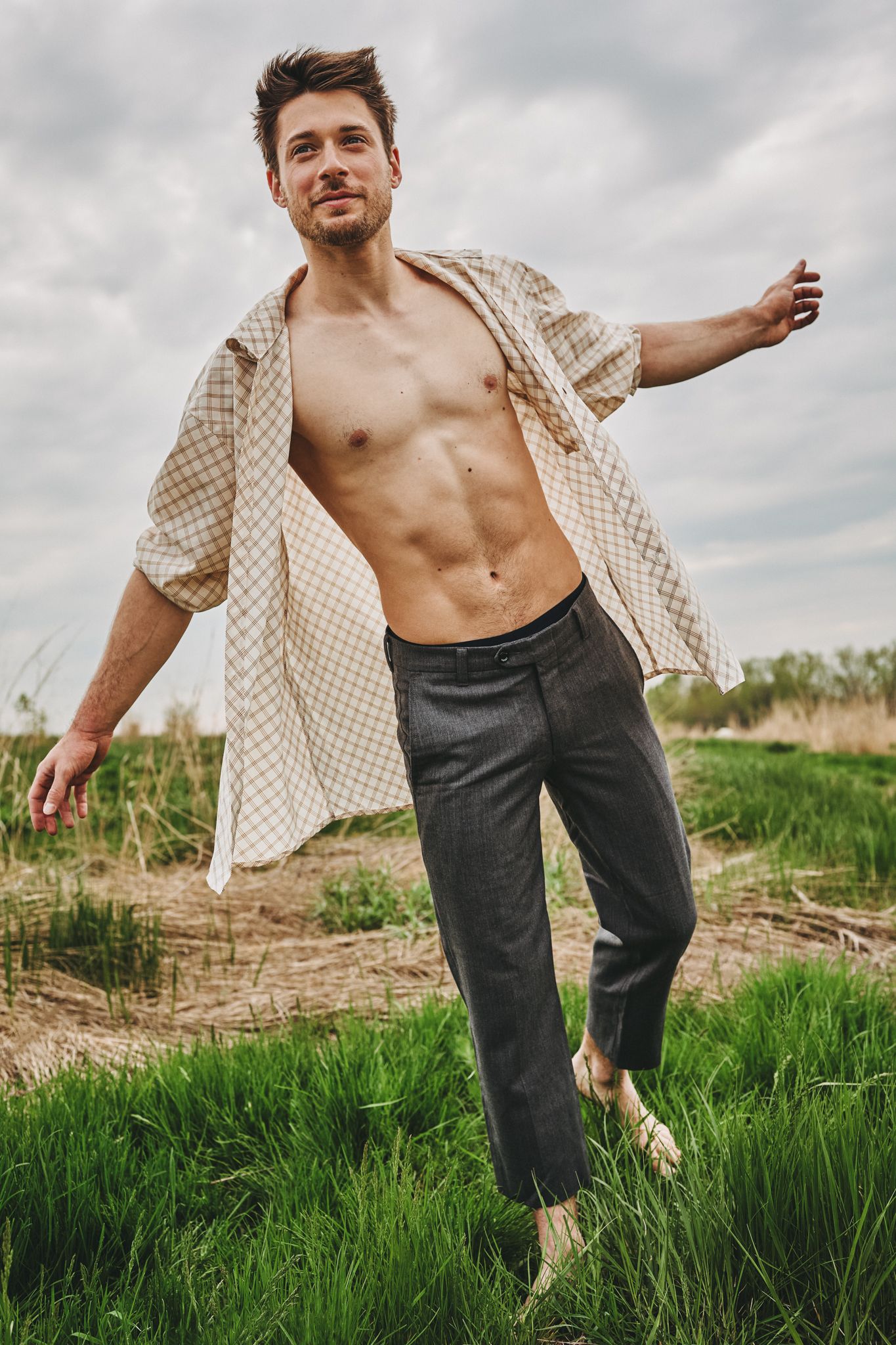 Person in checkered shirt and dark pants stands in grassy field against cloudy sky.