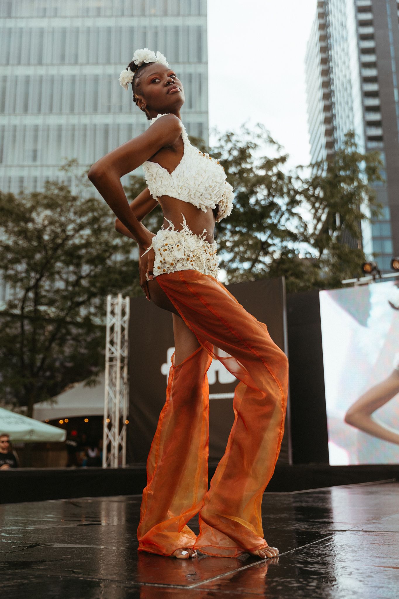 Performer in white lace crop top and orange bell bottoms strikes a pose on outdoor stage with city buildings in background.