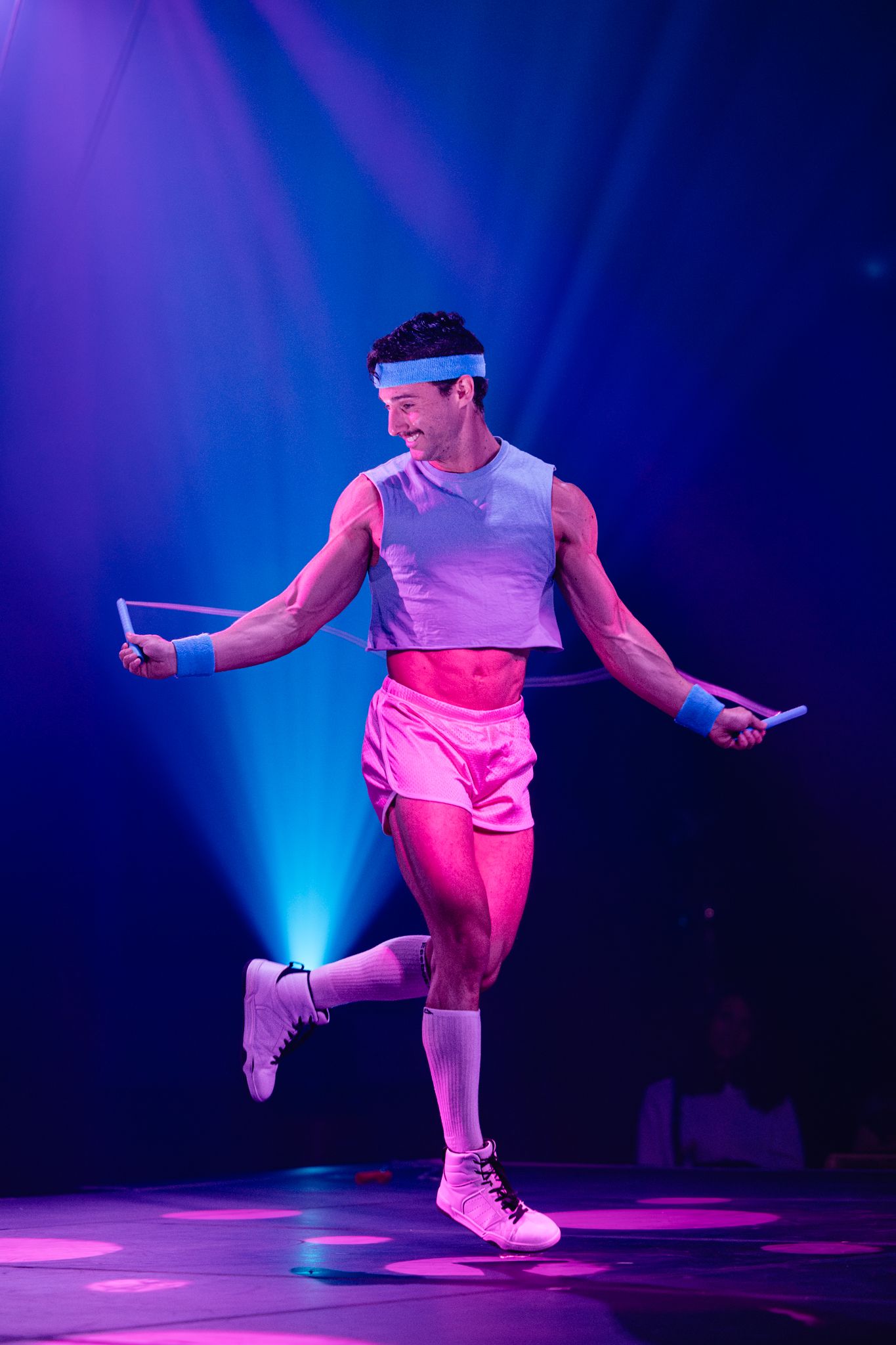 Dancer in white athletic wear performs energetic movements under blue and pink stage lighting.