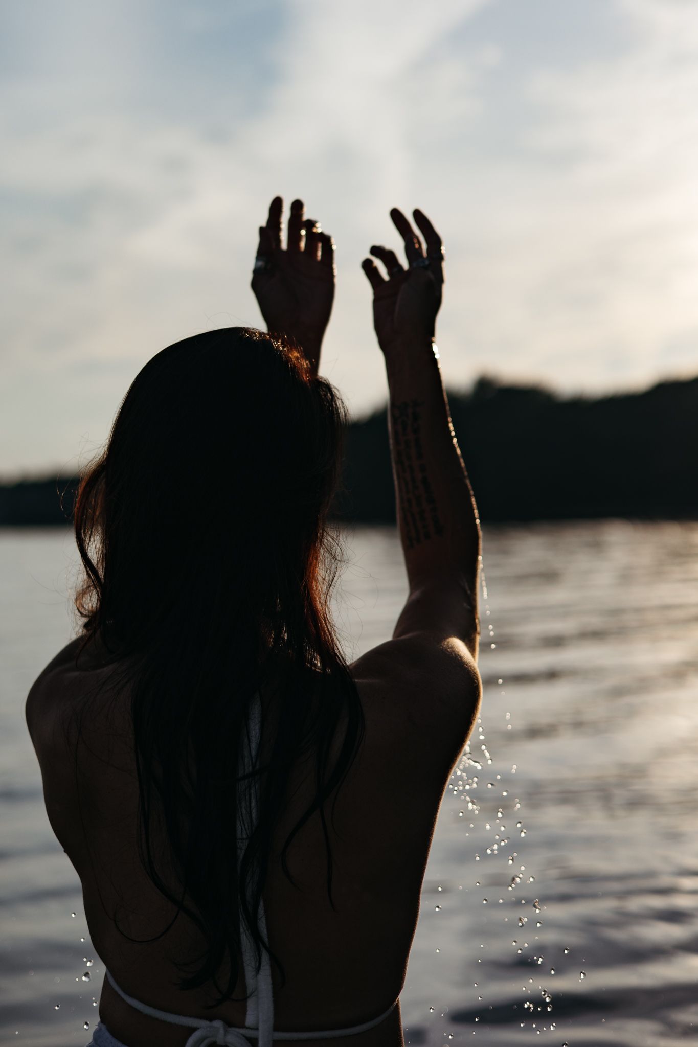 Silhouette of a person raising hands against a sunset at a lake, with water droplets splashing in the backlight.