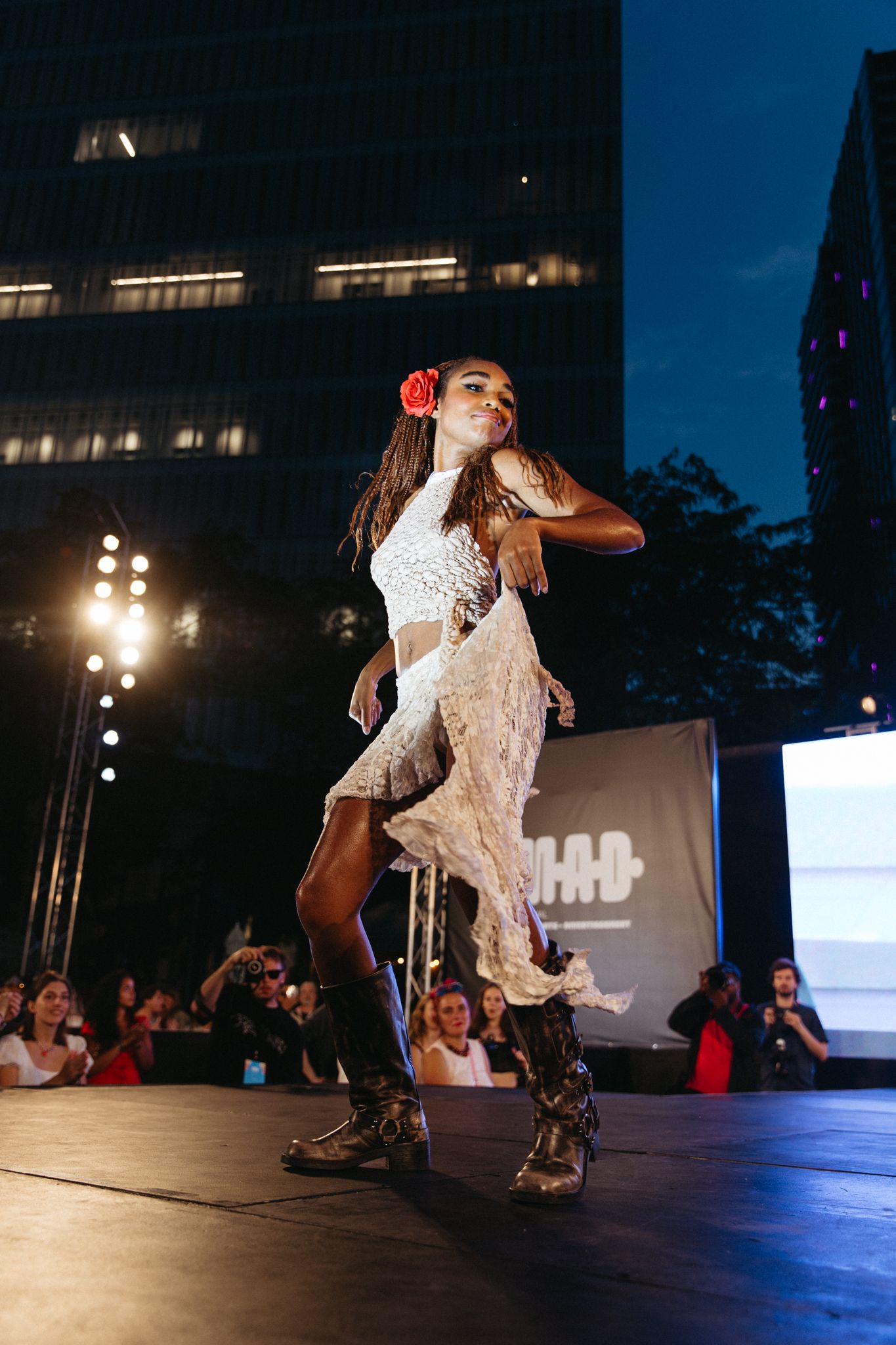 A performer in a white lace outfit and boots dances energetically on a stage at night with city buildings in the background.