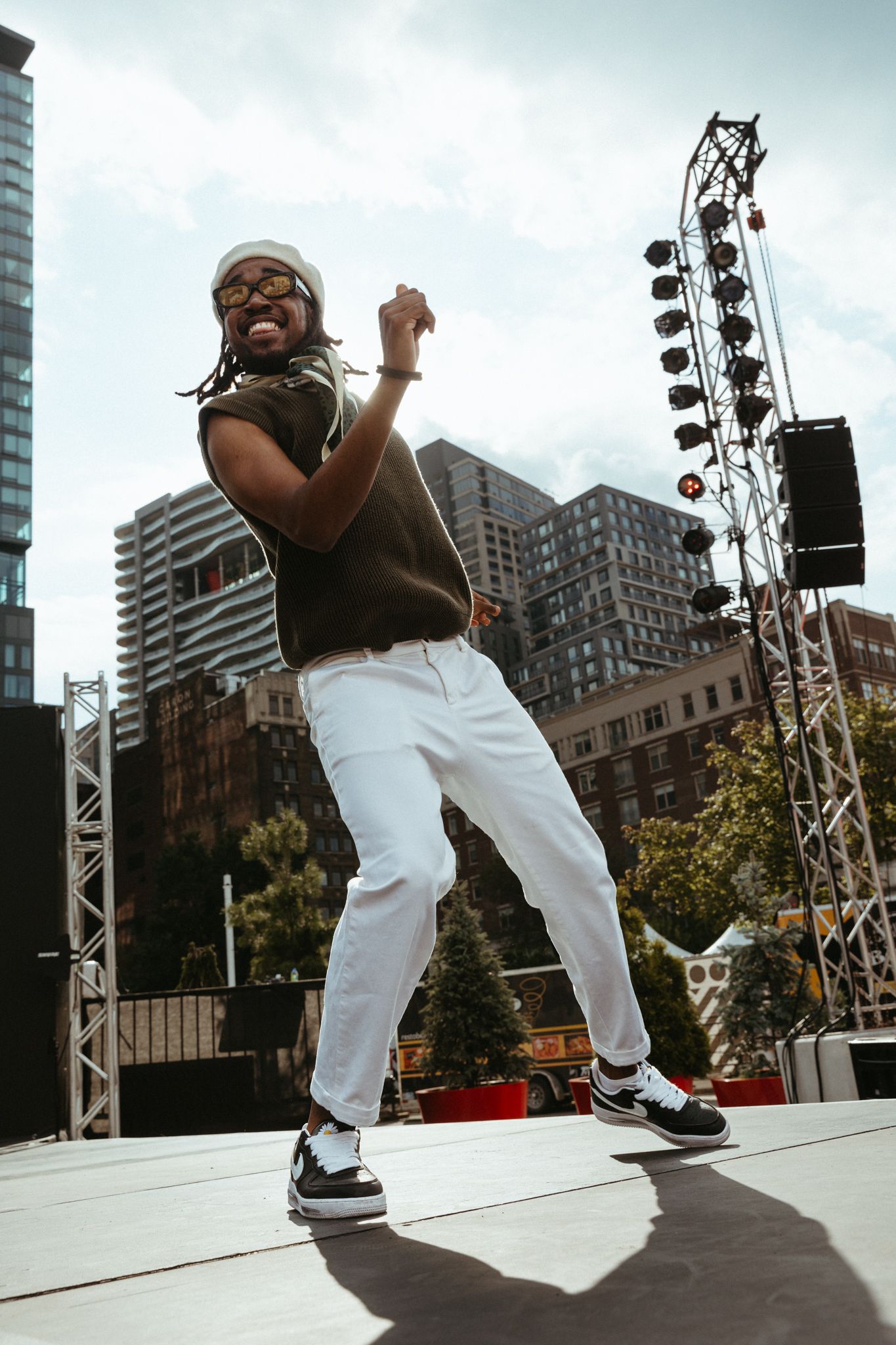Dynamic performer in white pants moves energetically across an outdoor stage with city buildings in the background.