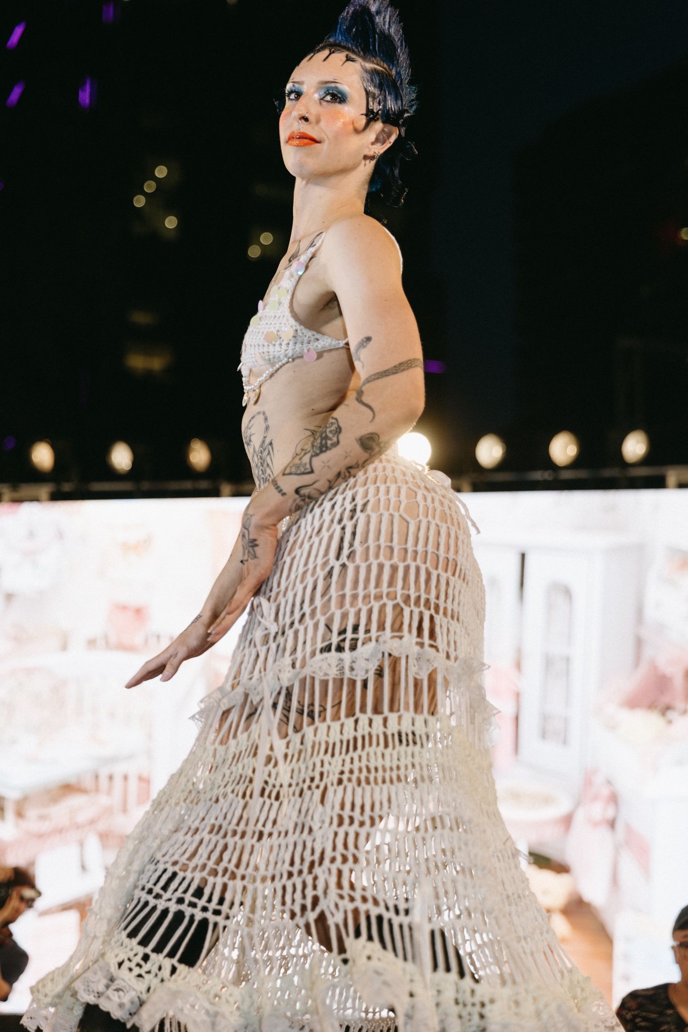 Model wearing an artistic white crochet dress with dramatic styling on a fashion runway stage.