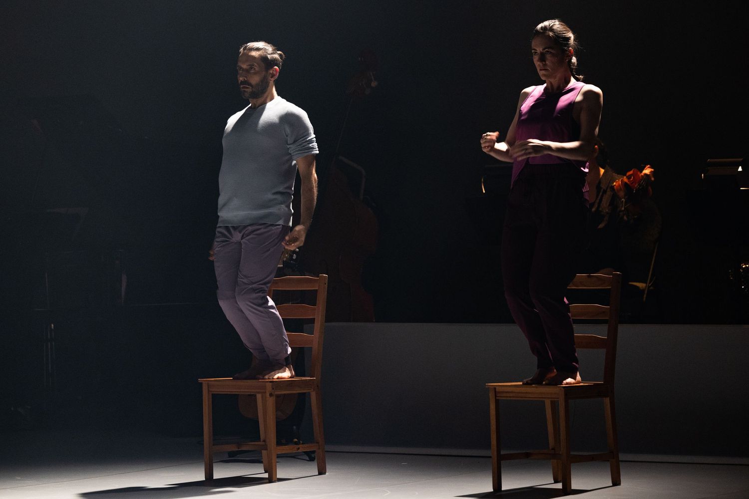 Two performers create dramatic tension in a dark theater scene with wooden chairs and dramatic lighting.
