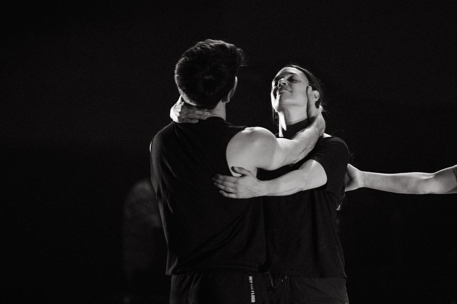 Two dancers in black outfits perform an intimate contemporary dance duet against a dark backdrop.
